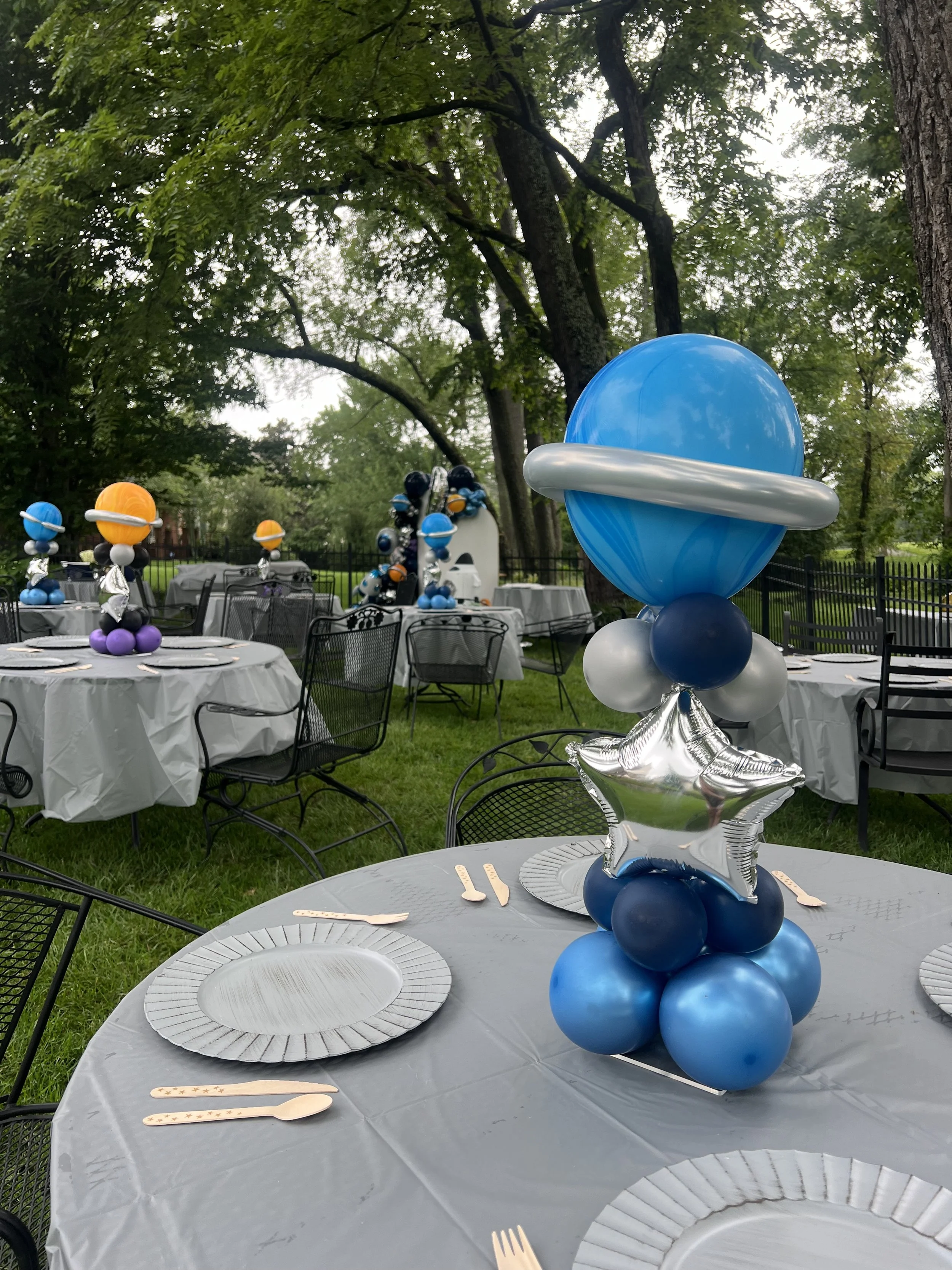 Outdoor party setup with space-themed balloon centerpieces on tables covered with gray tablecloths, surrounded by black chairs, in a park with green trees and a black fence in the background.