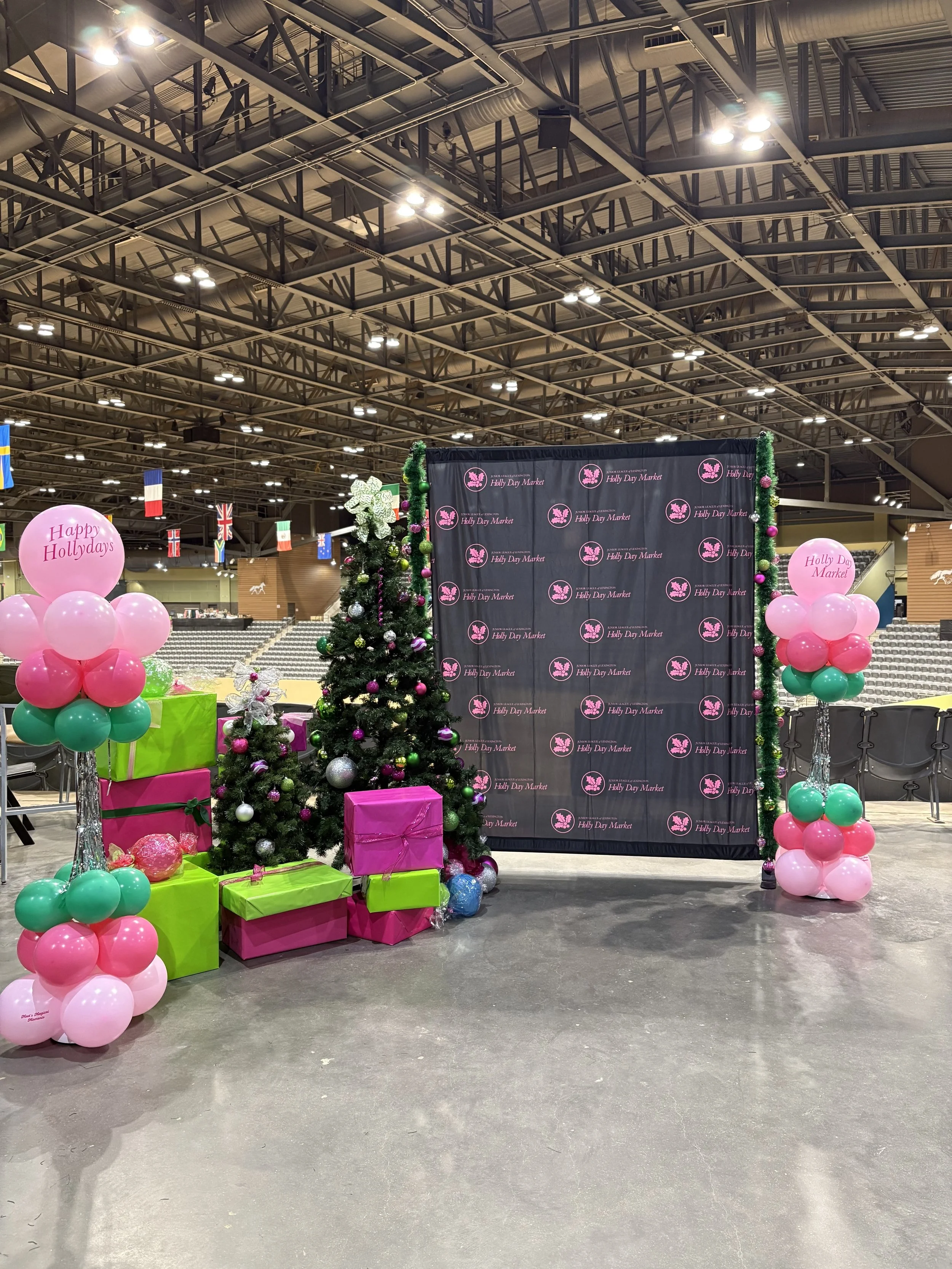 Christmas setup with decorated trees, wrapped presents, balloon displays, and a backdrop with 'Holly Days Market' repeated on it.