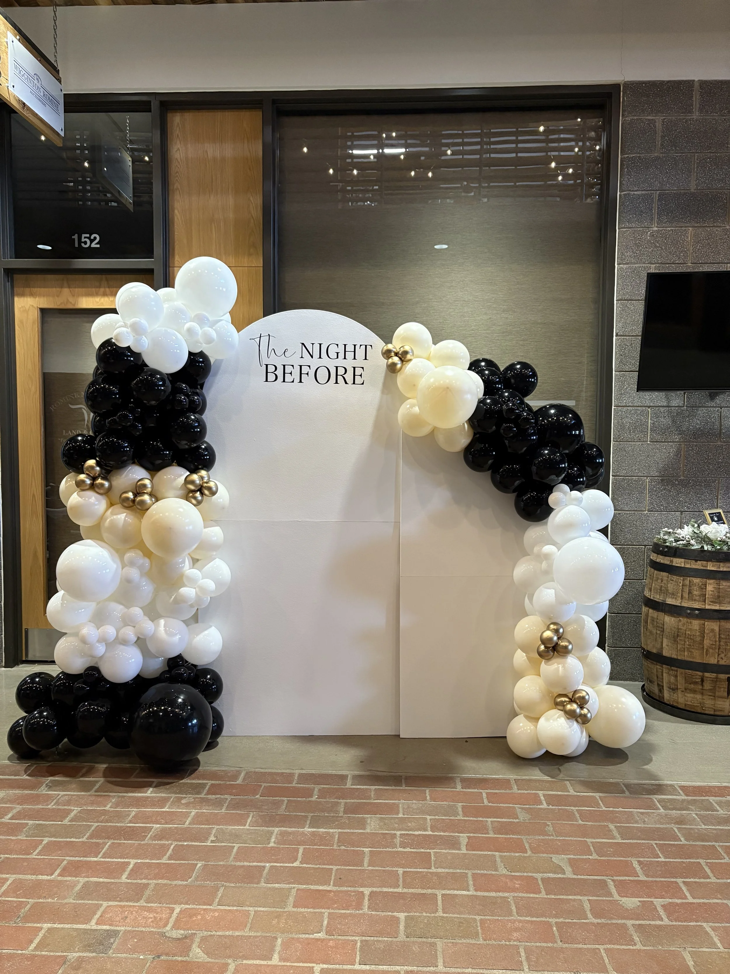 Decorative balloon setup with black, white, and gold balloons around a white backdrop with the words 'The Night Before' printed on it, likely for a celebration or event.