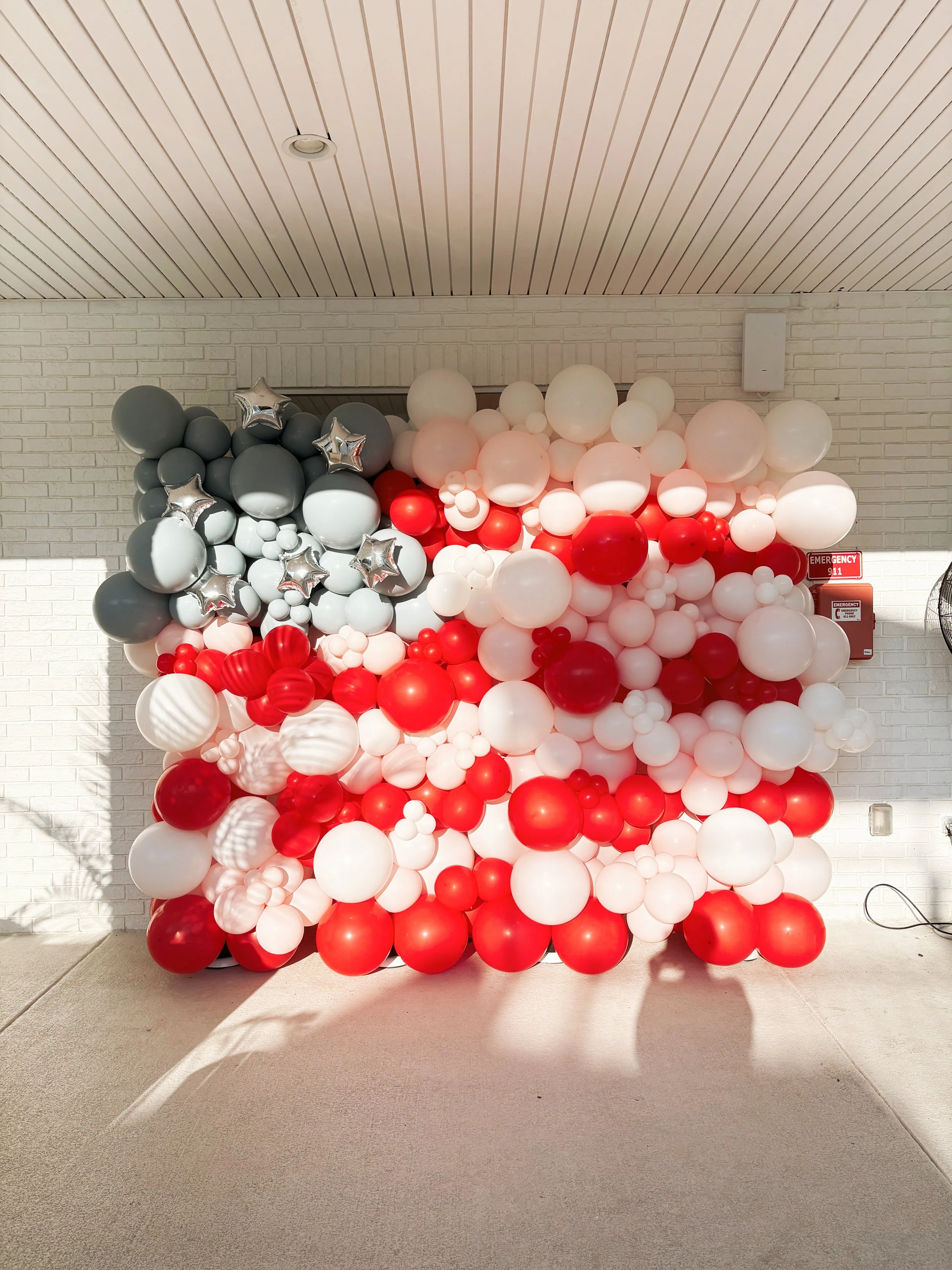 A large decorative balloon wall with gray, red, pink, and white balloons, some with star patterns, arranged in a cluster inside a home.