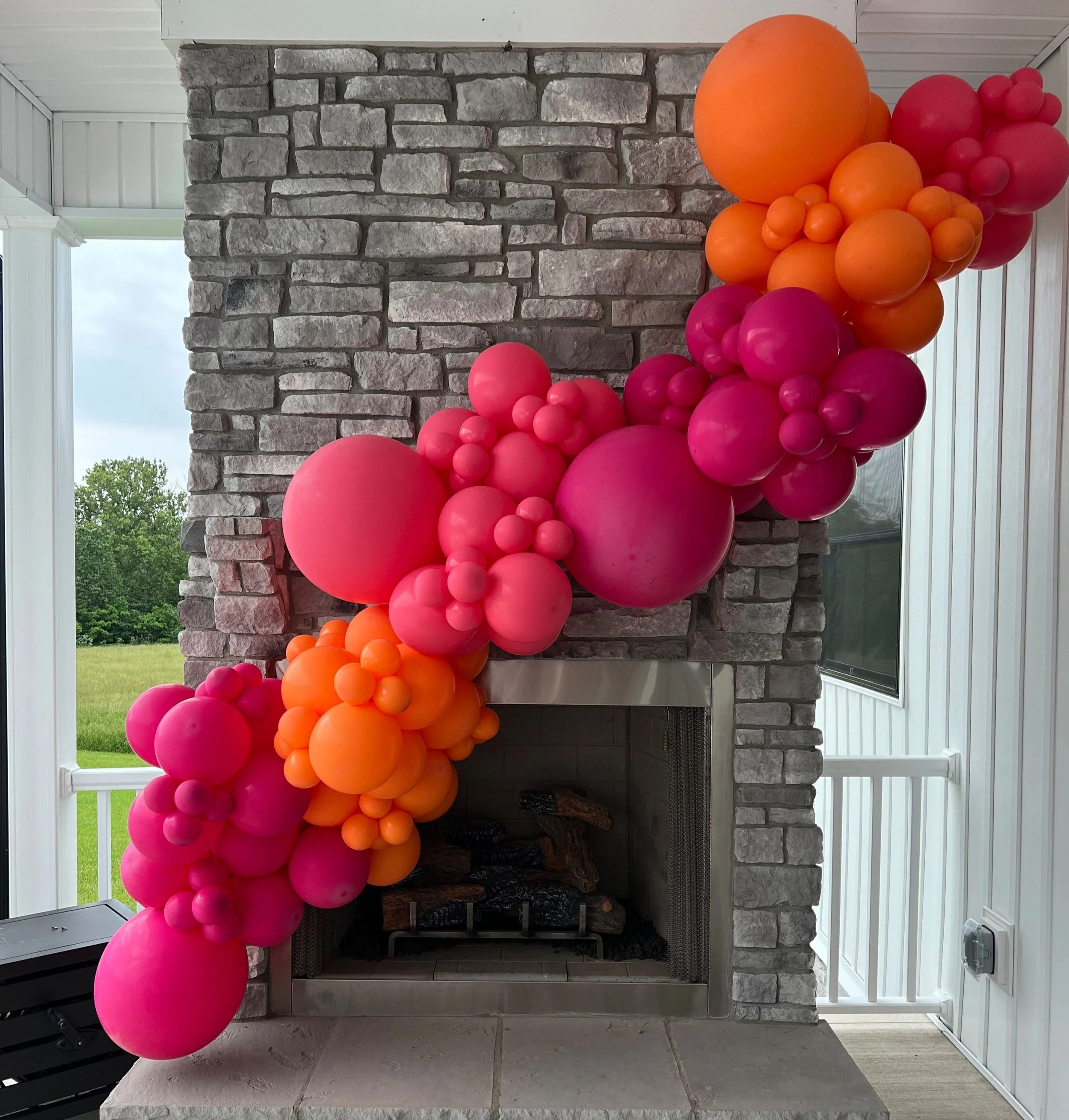 Colorful balloon garland in shades of pink and orange draped over a gray brick fireplace.