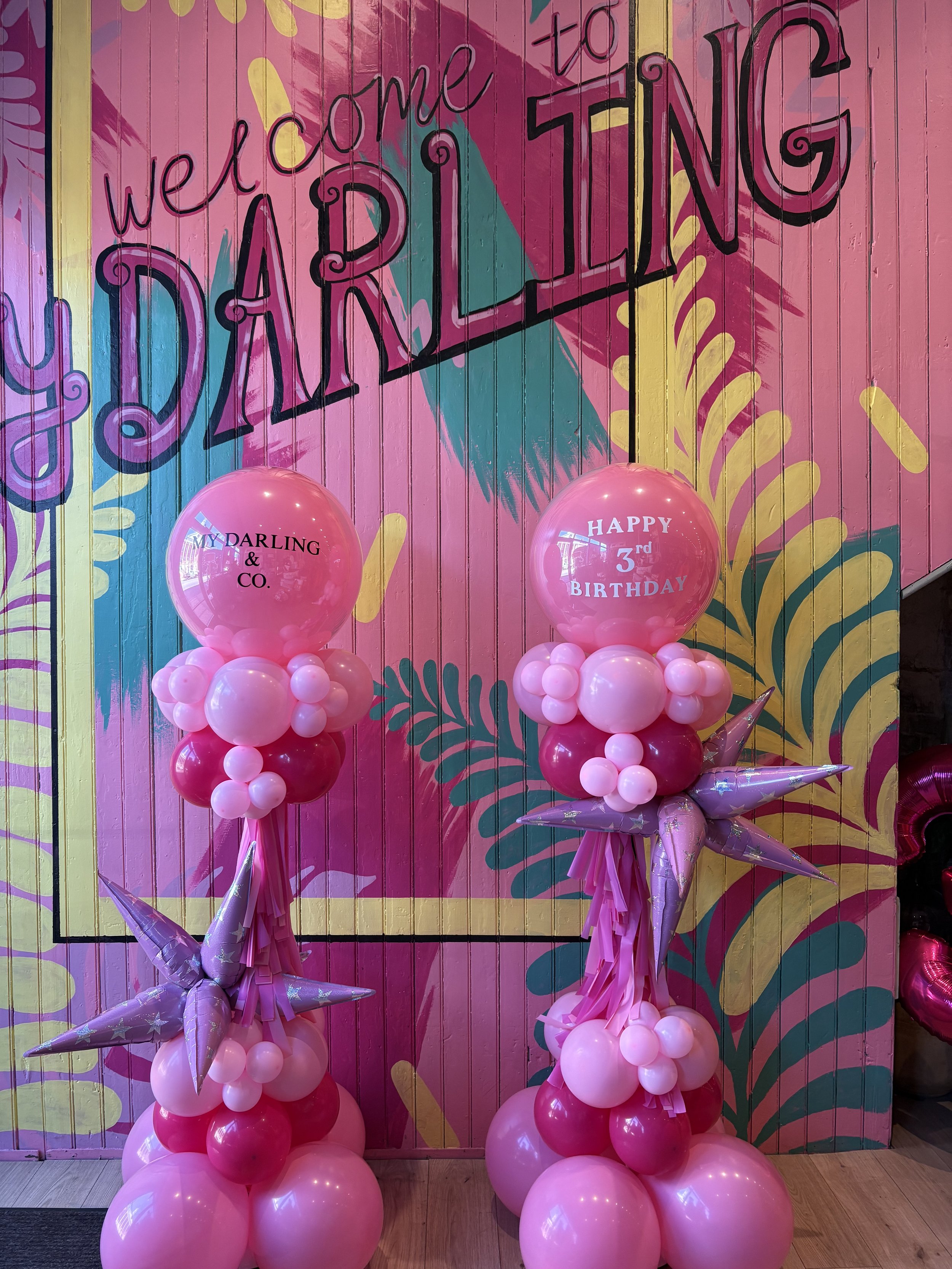 Christmas setup with decorated trees, wrapped presents, balloon displays, and a backdrop with 'Holly Days Market' repeated on it.