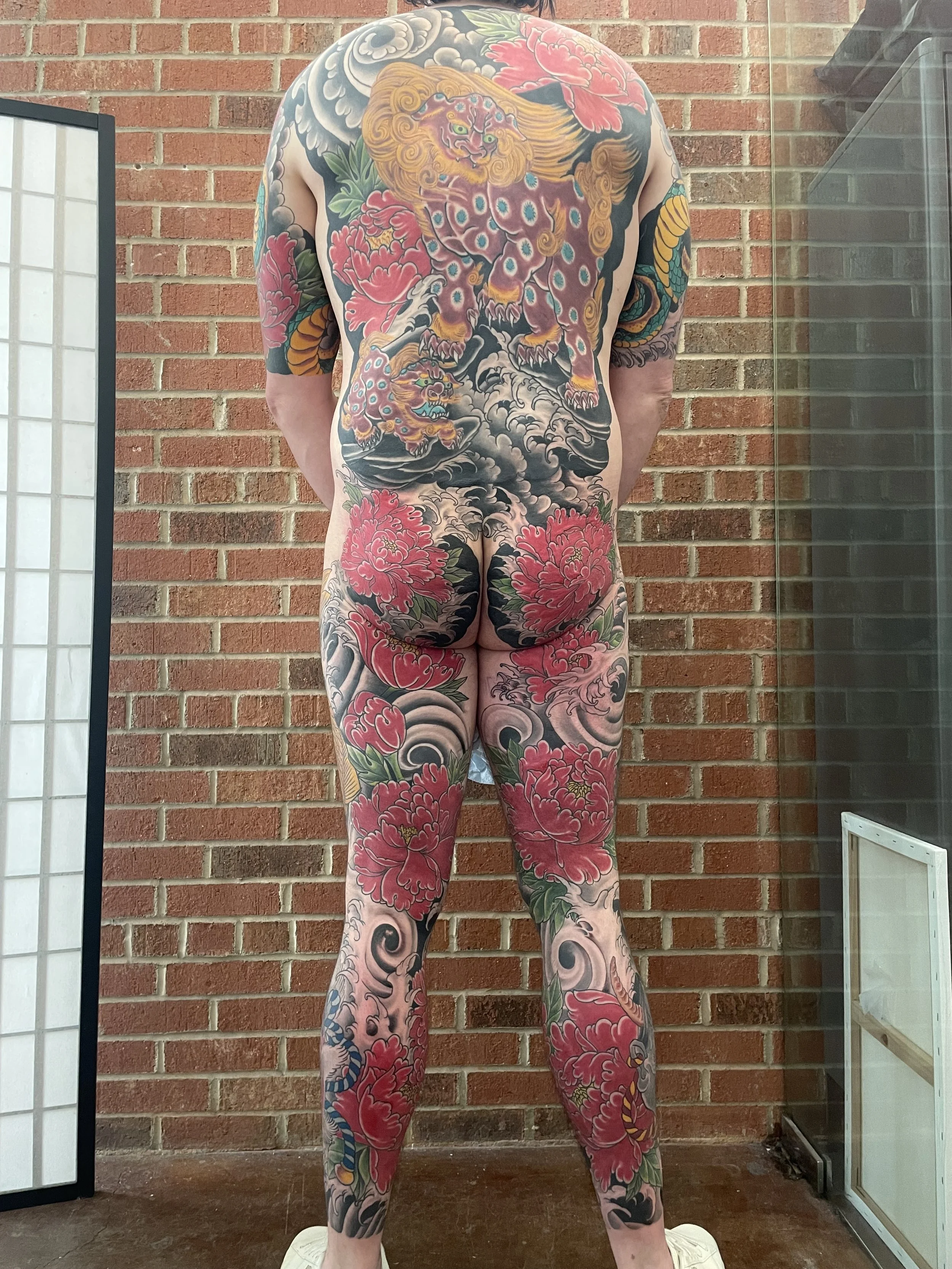 A person with full-body tattoos of Japanese-style motifs, including a large Foo Dog on their back, surrounded by colorful peonies and clouds, standing against a brick wall.