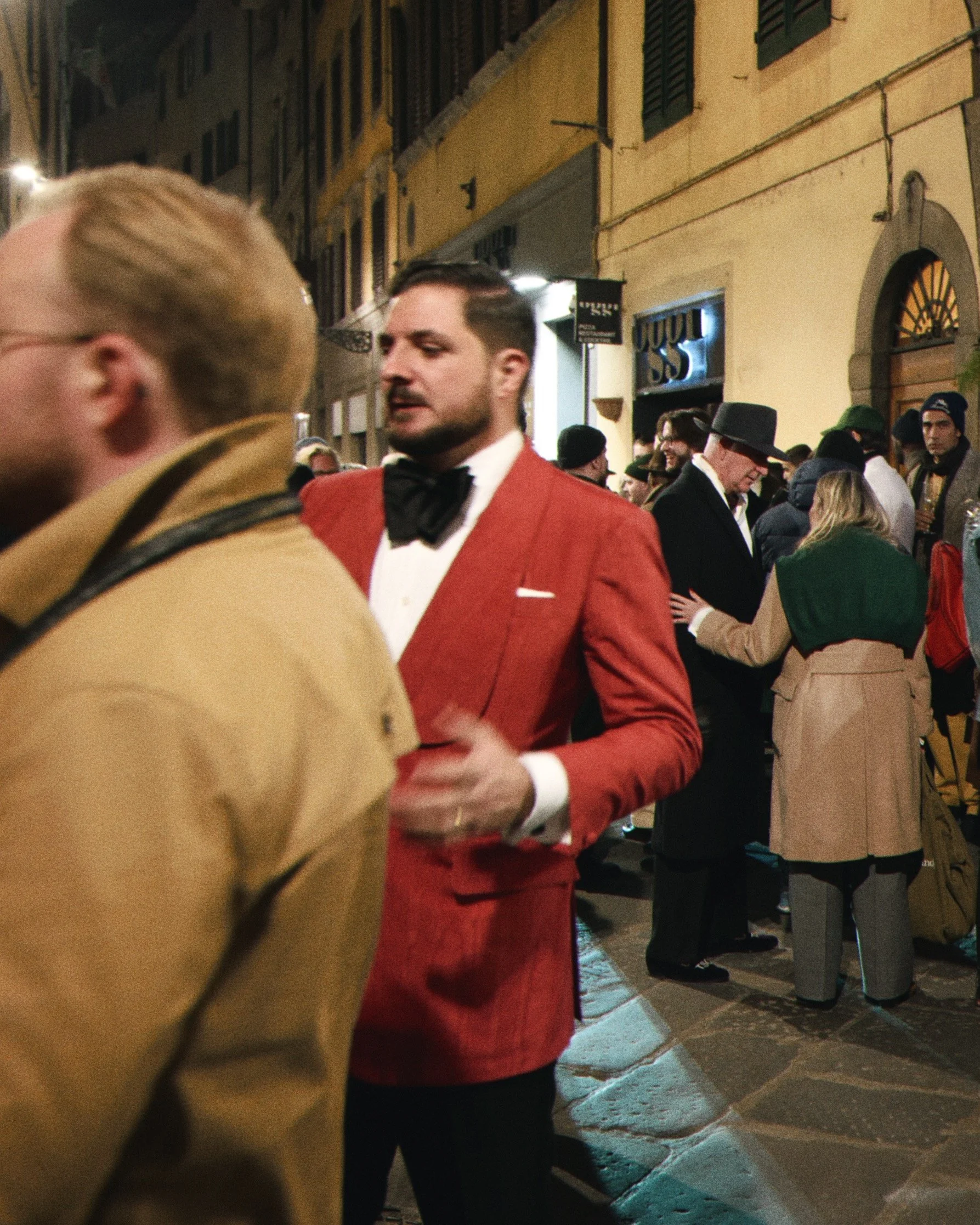 People walking on a street at night, with some dressed in formal suits and coats, in what appears to be a city with old architecture and cobblestone streets.