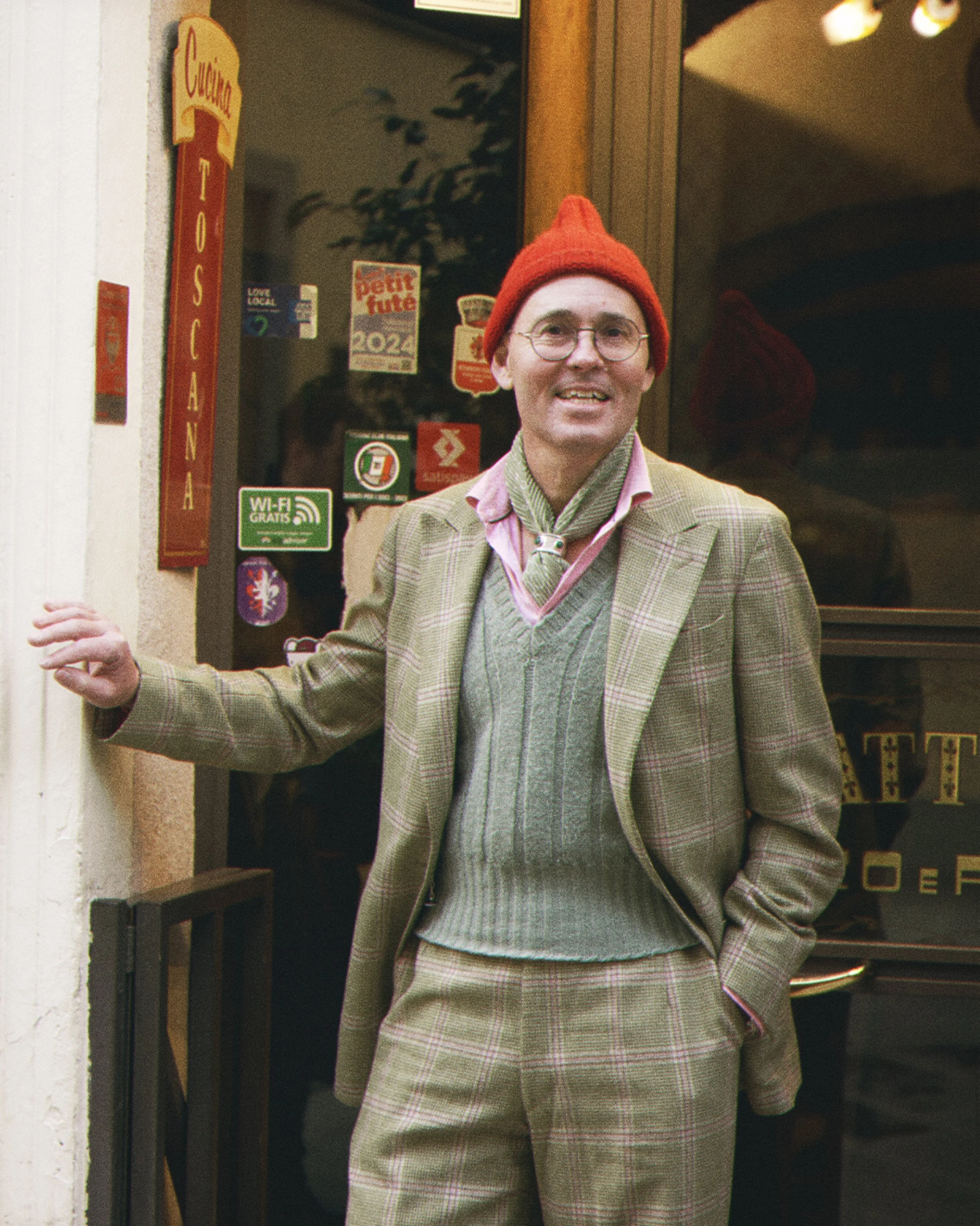 A person with glasses wearing a red knit hat, a beige plaid suit, and a green scarf, standing outside a store.