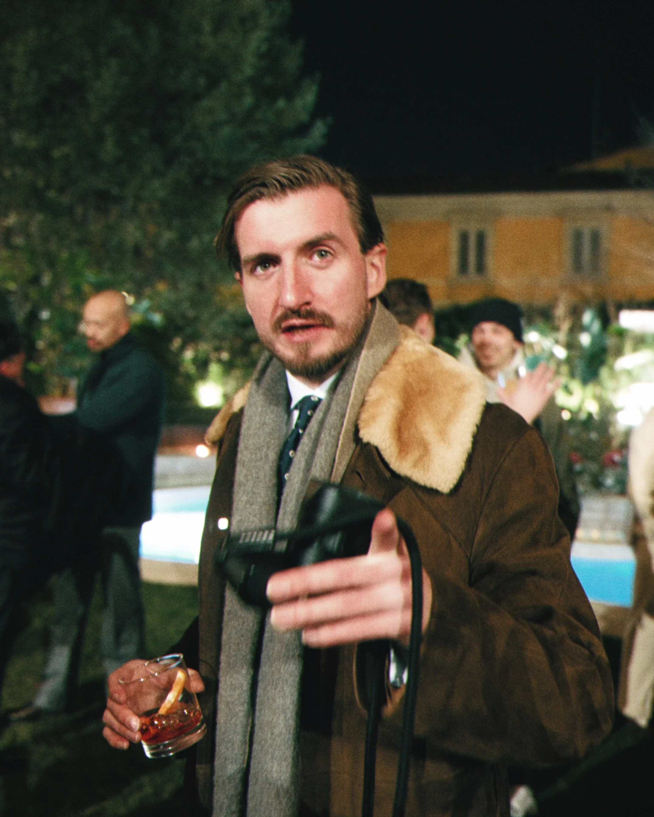A man wearing a brown jacket with a fur collar, holding a glass of dark liquid, at an outdoor gathering at night with other people in the background.