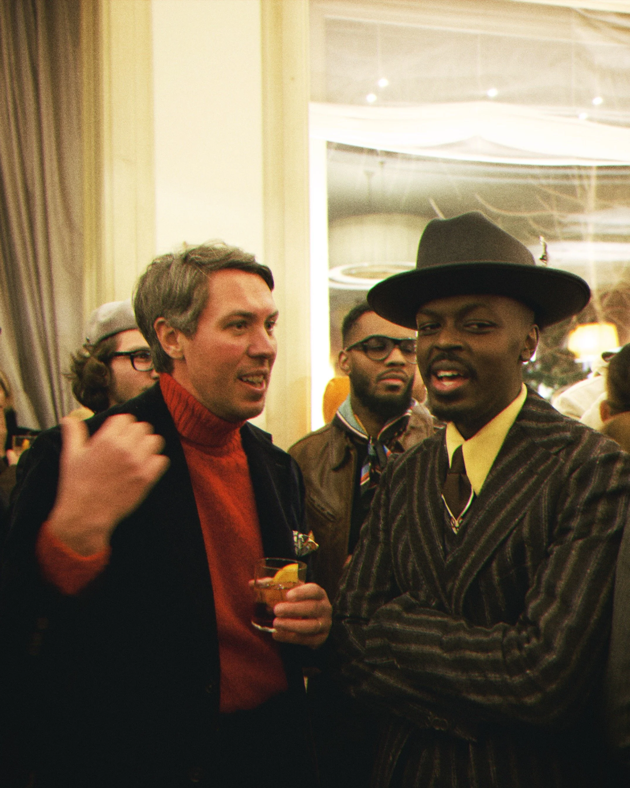 A group of people seen at a social gathering, with a woman in a red turtleneck and black jacket talking to a man wearing a hat and a striped blazer, holding a glass of drink, and with other individuals in the background.