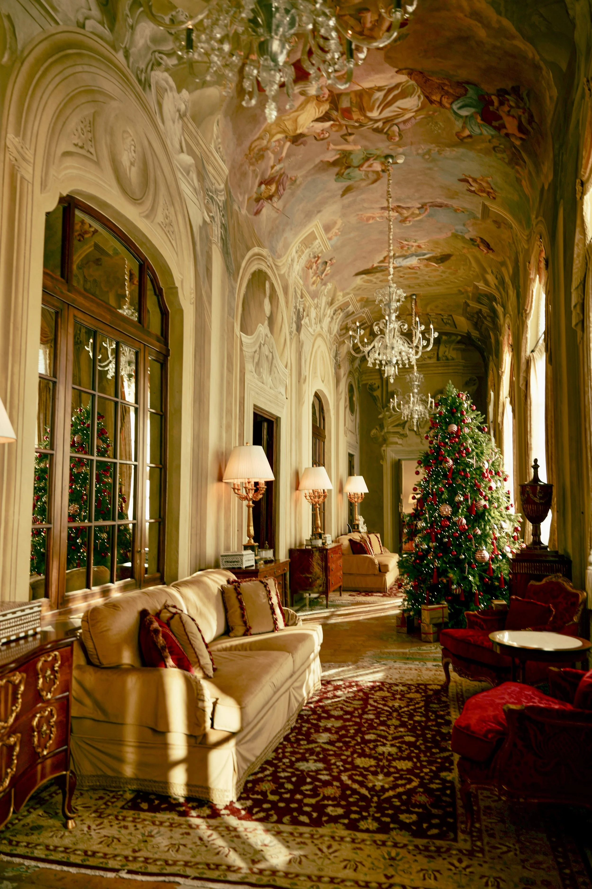 A luxurious living room decorated for Christmas with a decorated Christmas tree, a chandelier, plush seating, ornate curtains, large windows, and a painted high ceiling depicting an elaborate scene.