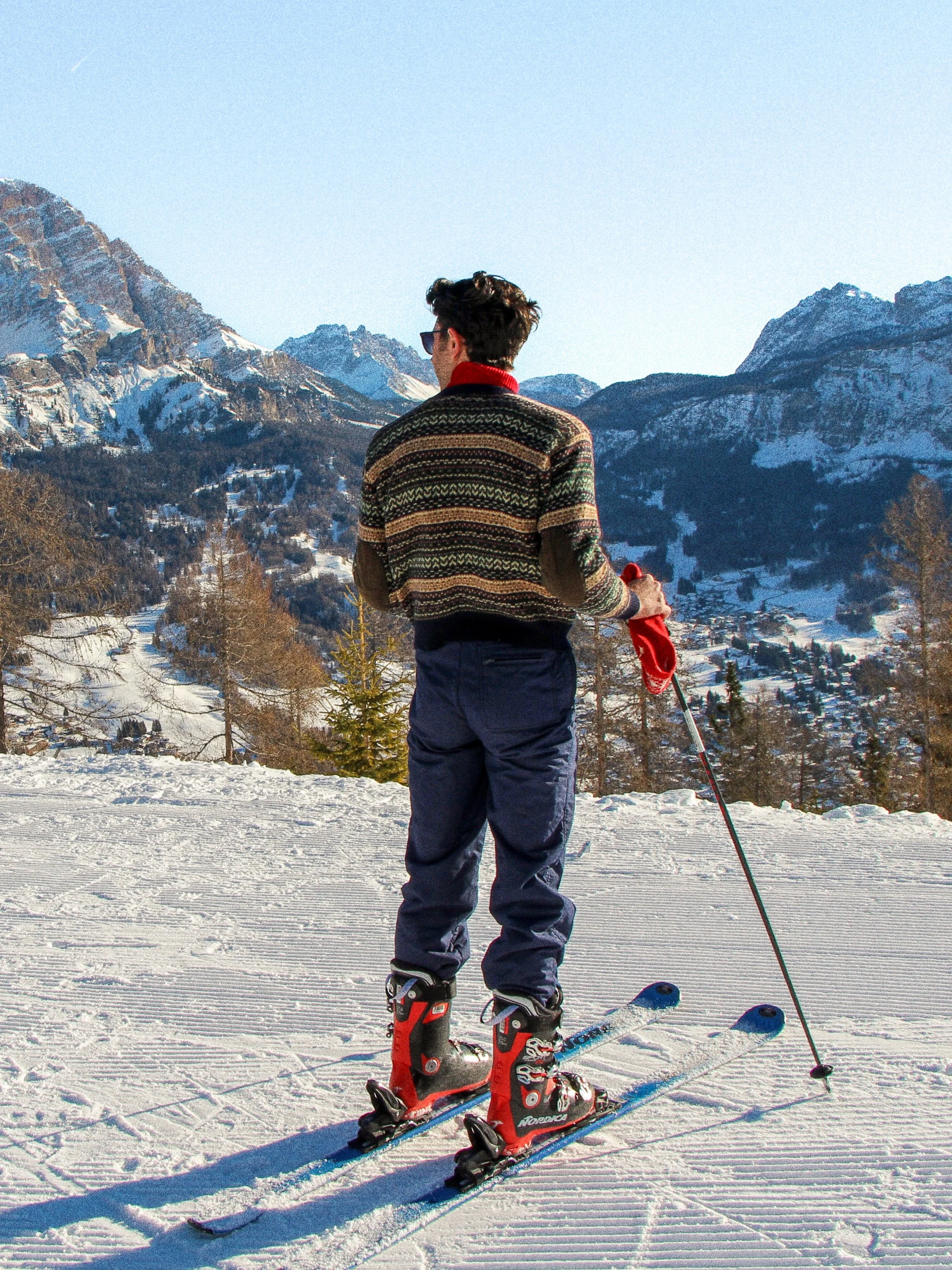 A person in colorful winter sweater and ski gear standing on skis with ski poles in a snowy mountain landscape.