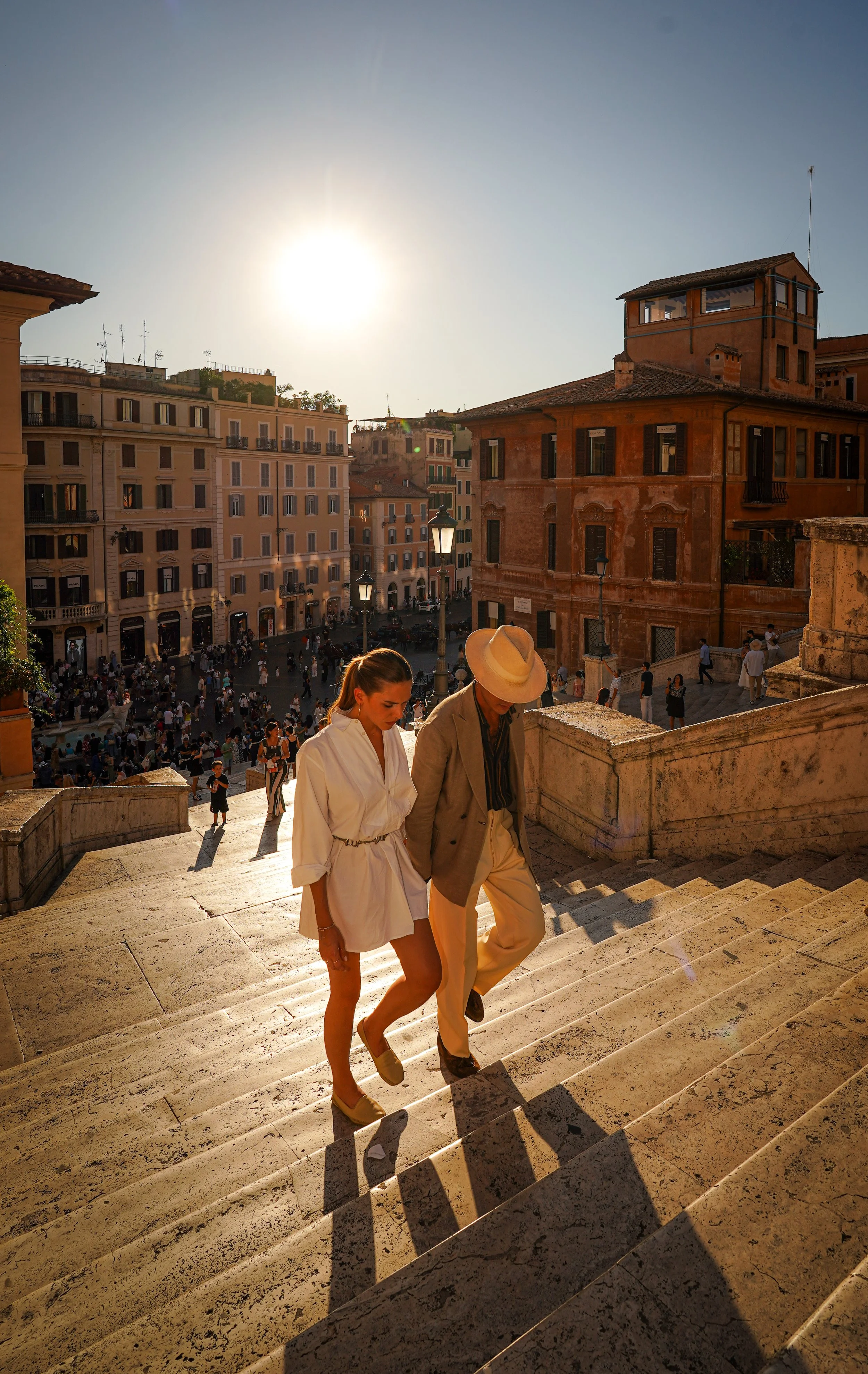 Two people walking up stone stairs in a European city square during sunset. The woman wears a white dress and the man wears a suit with a fedora hat. The background features historic buildings and a crowded square.