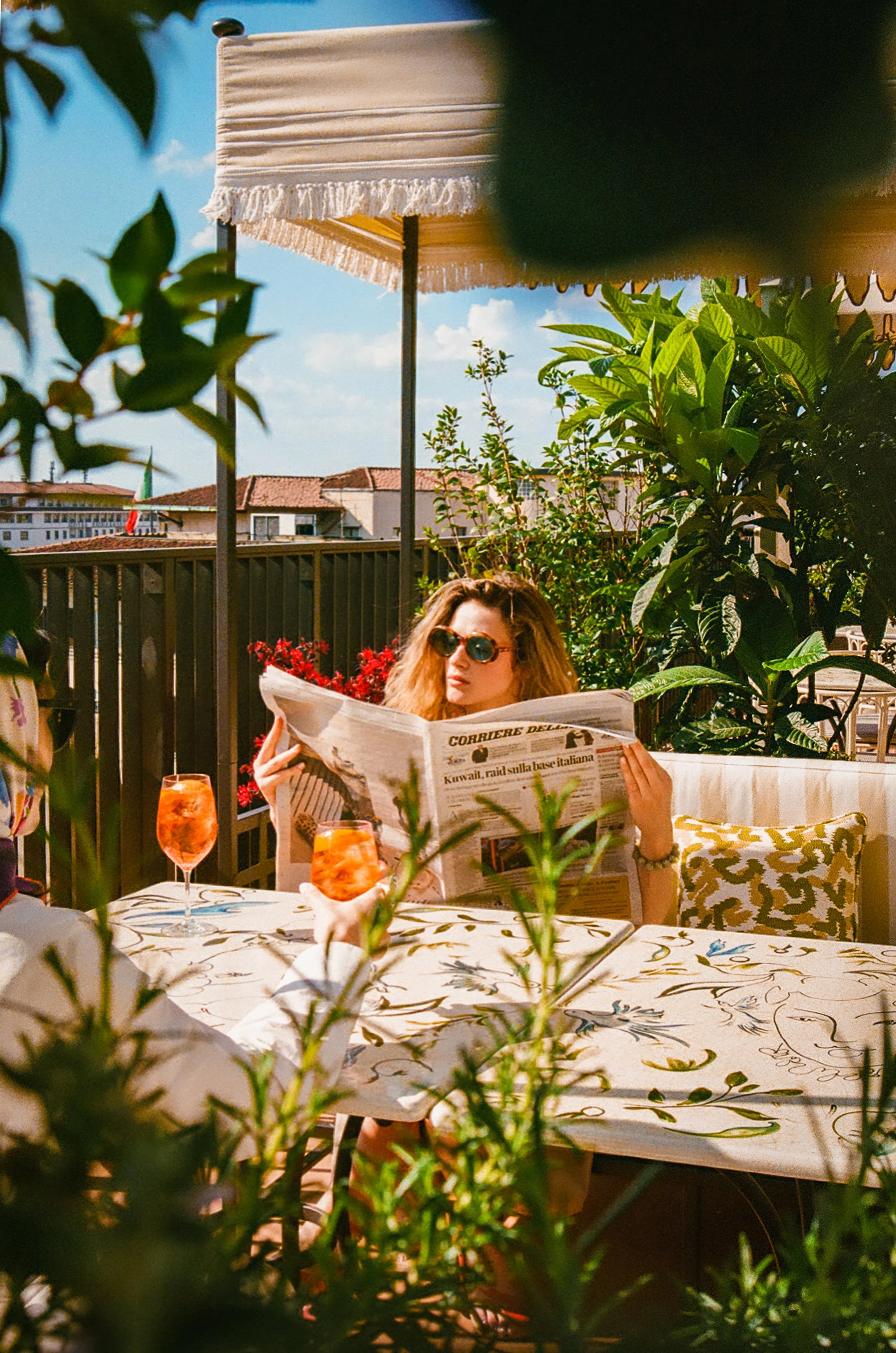 A woman with curly hair and sunglasses reading a newspaper at an outdoor dining table with floral tablecloth and two glasses of orange drink, surrounded by greenery and plants, on a sunny day.