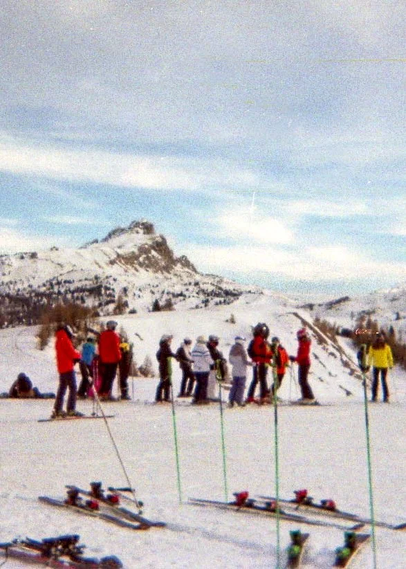 Group of people in winter clothing on a snowy mountain slope, with ski equipment lying in the snow in the foreground, and a mountain with snow-covered peaks in the background.