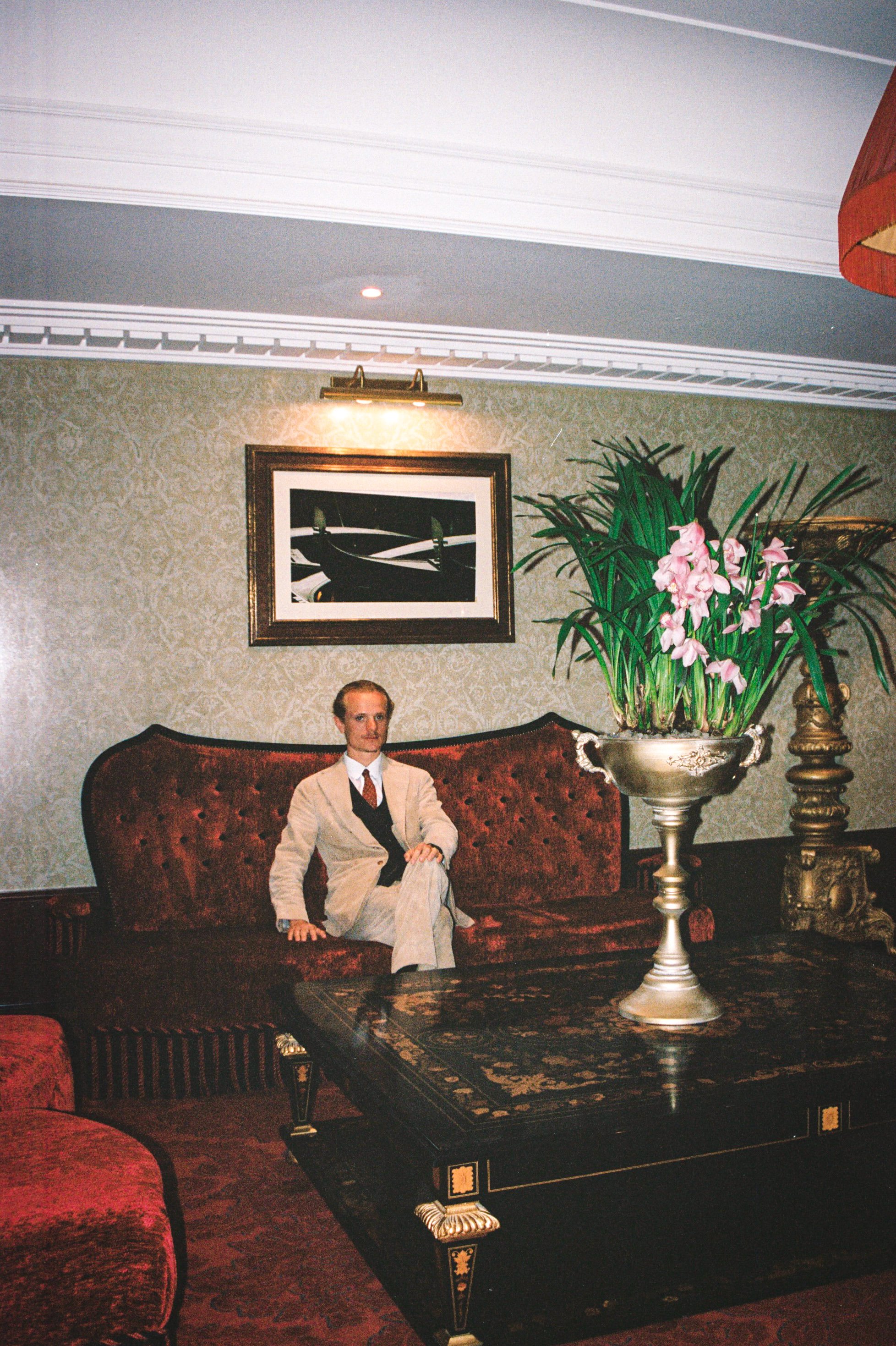 Man sitting on a red velvet sofa in a living room with green wallpaper, a large potted plant with pink flowers on a black table, framed artwork, and ornate furniture and decor.