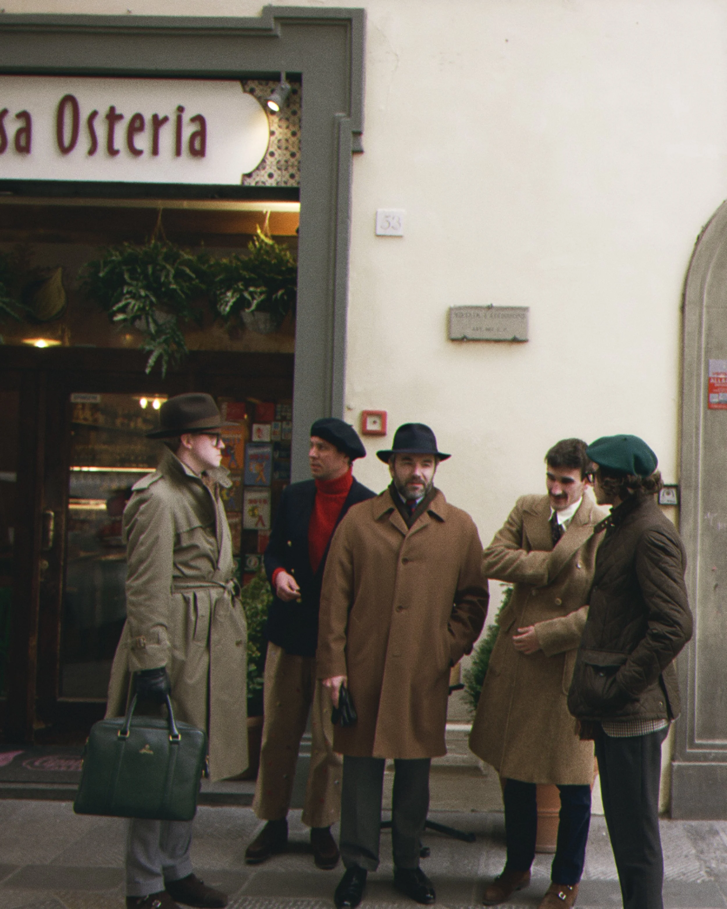 A group of people in vintage clothing, standing outside a shop entrance with a sign that reads "Osteria." Several of the individuals are wearing trench coats, hats, and gloves, suggesting a mid-20th-century time period. They appear to be engaged in c
