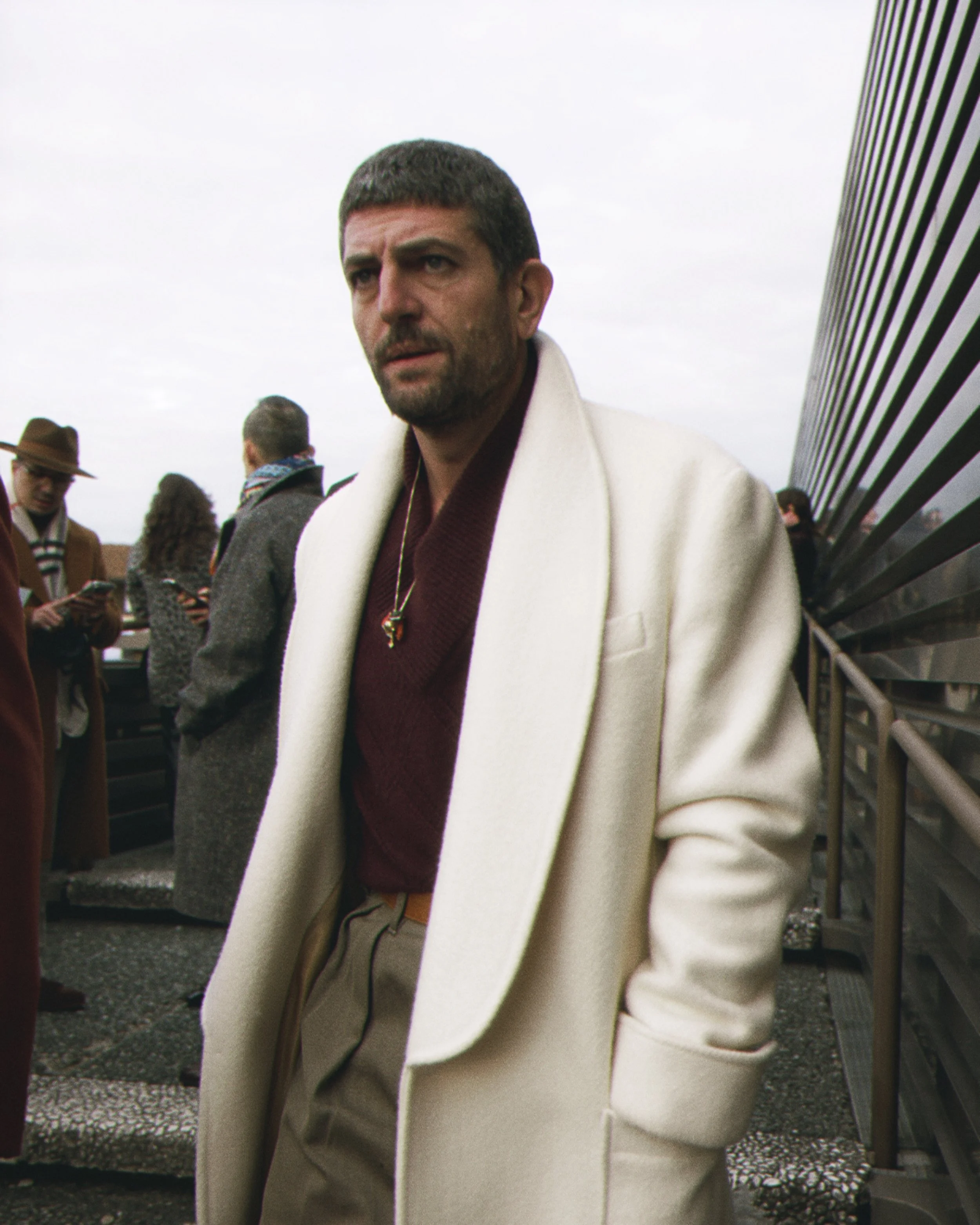 A man with short dark hair and a beard in a white coat standing outdoors with other people in the background, including a person with a brown hat and glasses, in an overcast setting with a modern building with horizontal lines on the right.