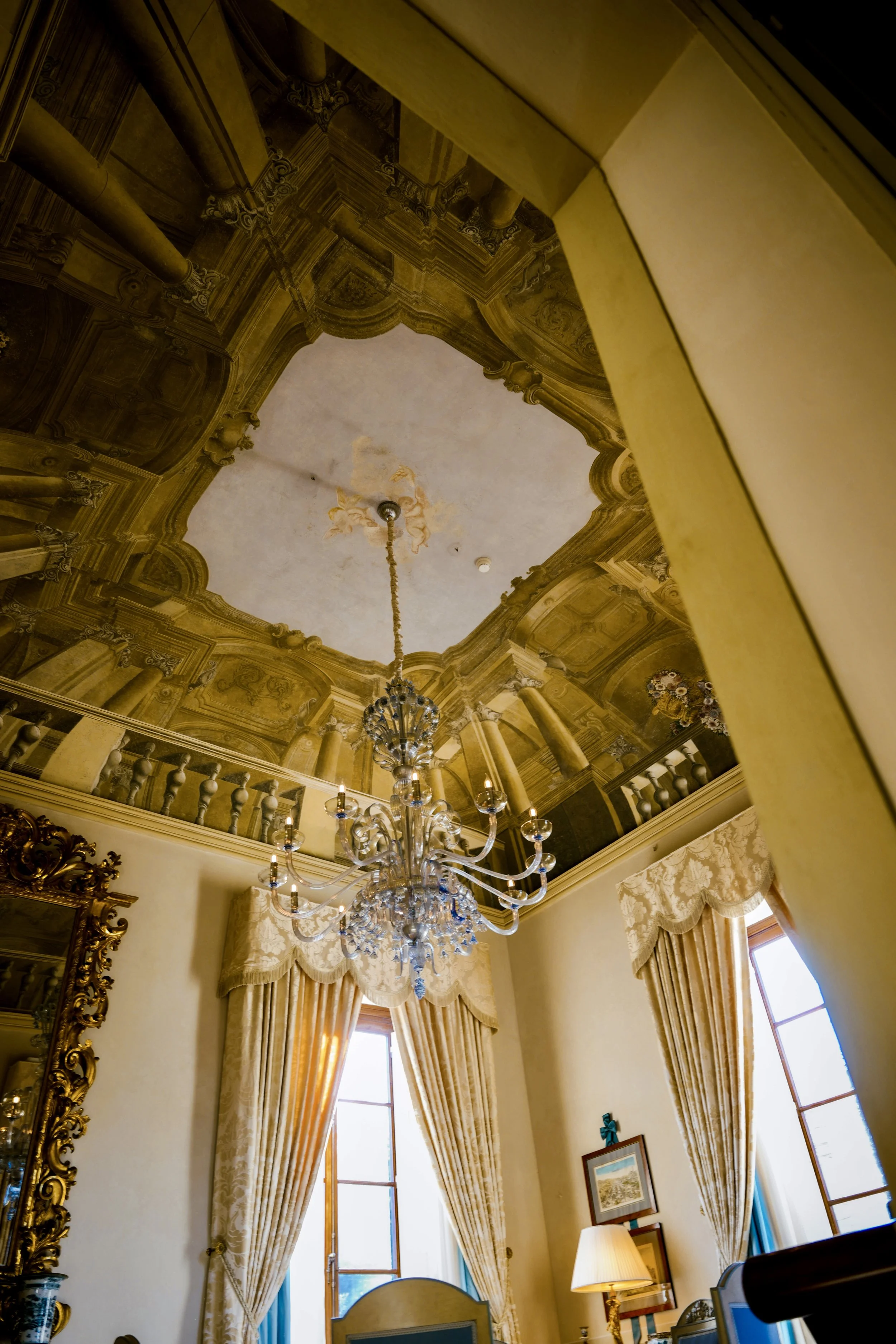 Elegant hotel room with ornate ceiling, chandelier, tall windows with cream curtains, and classical decor.