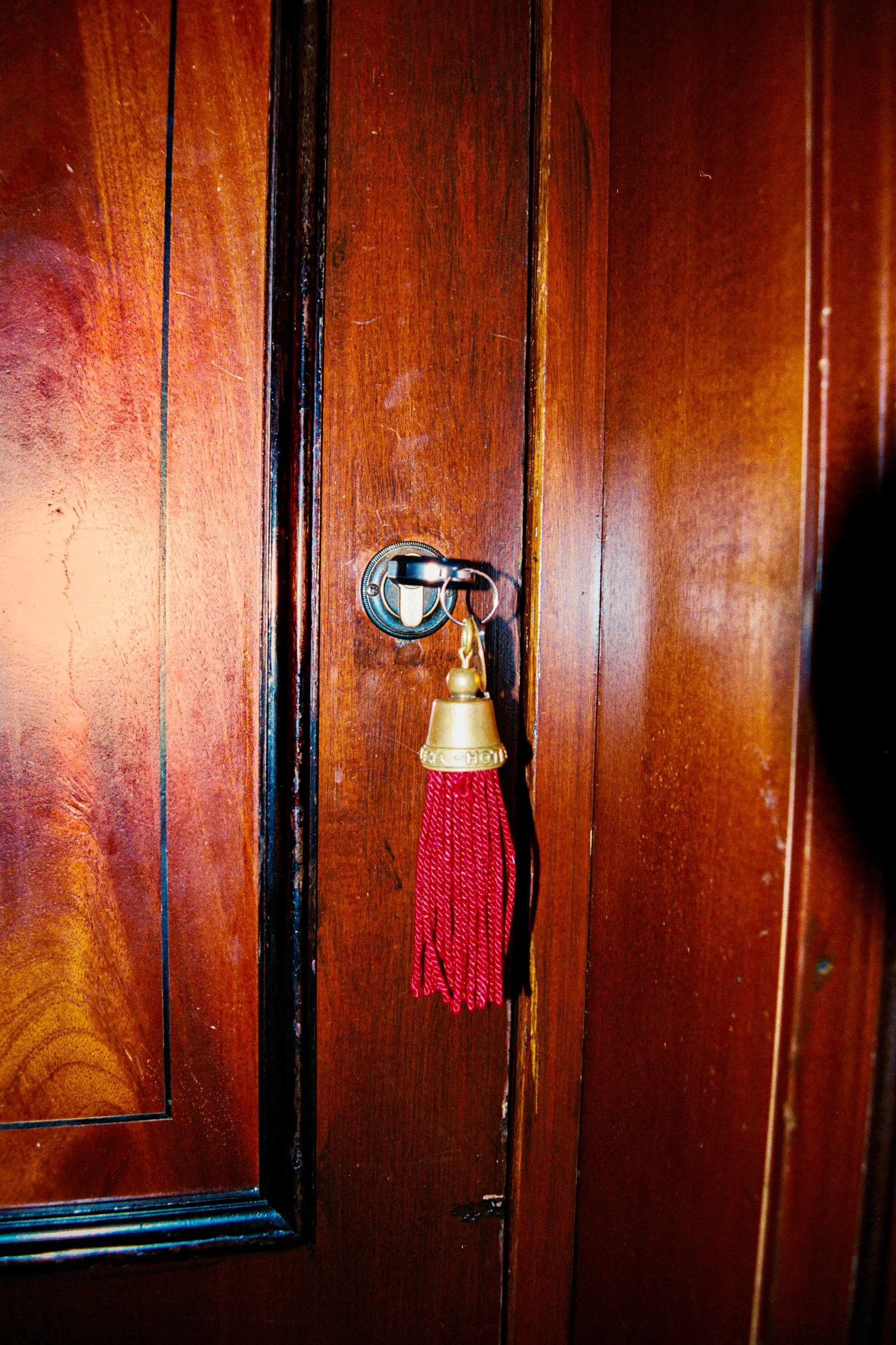 Wooden door with a keyring hanging from a silver door hook. The keyring has a large, gold-colored bell with a red tassel attached.