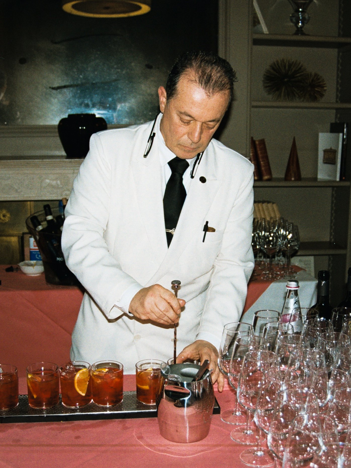 A man in a white coat and black tie preparing drinks at a bar or event setting, with various glasses of drinks and empty glasses on a table in front of him.