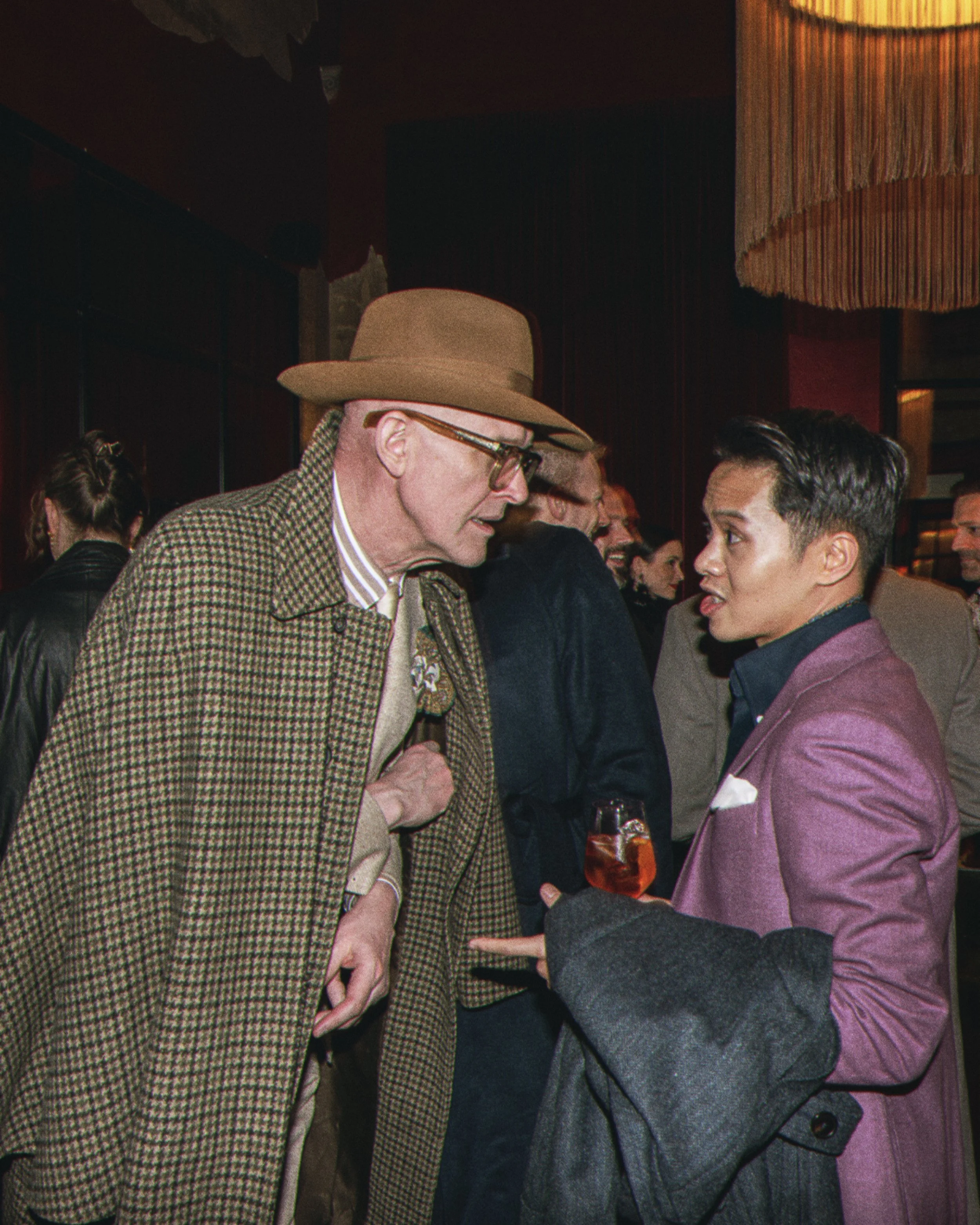 An older man wearing a brown hat, glasses, and a brown checkered coat, talking to a young man in a pink blazer at a social gathering.