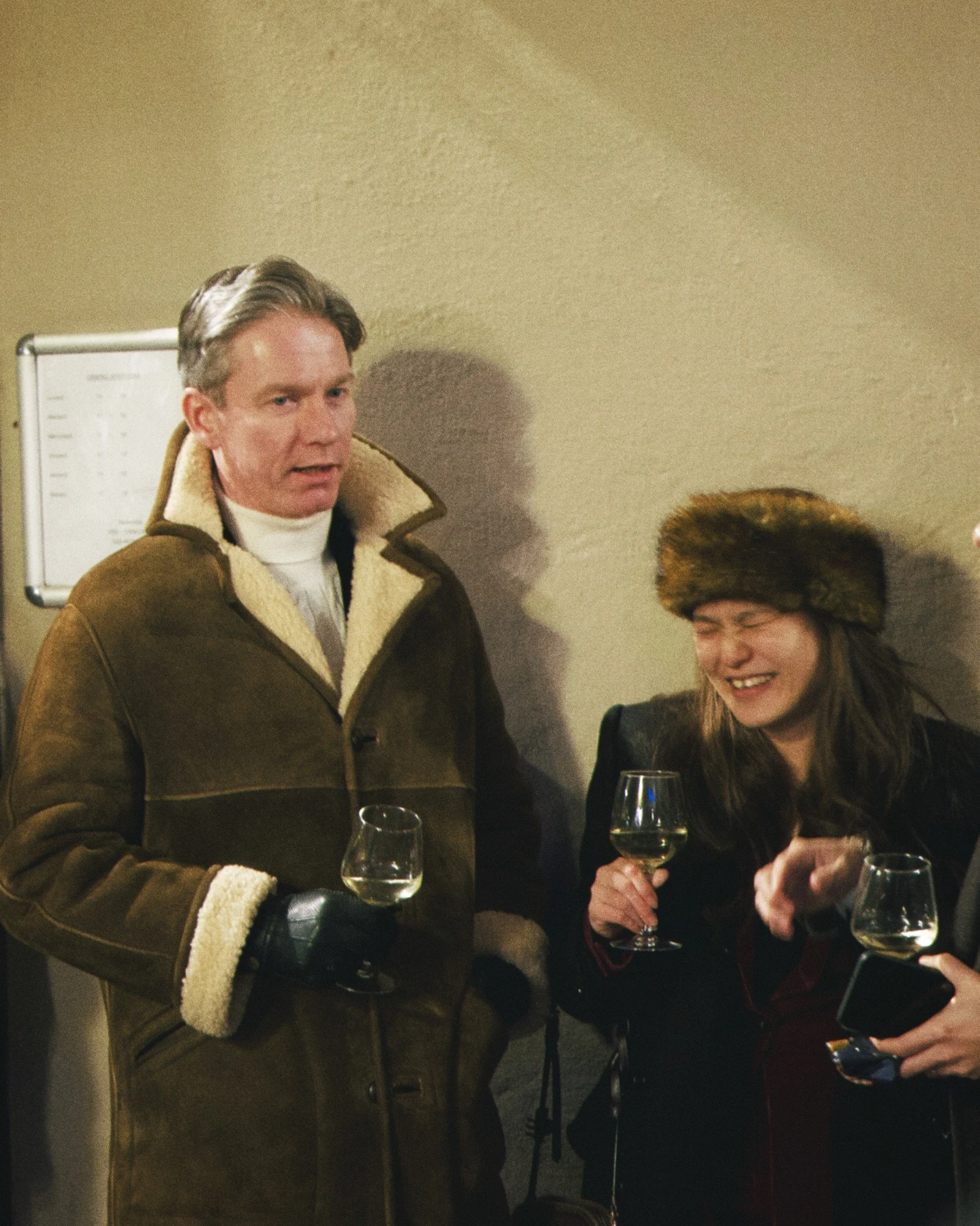 Two people at a social gathering, one man standing and one woman sitting, both holding glasses of white wine, with a neutral background and a whiteboard partly visible on the left.