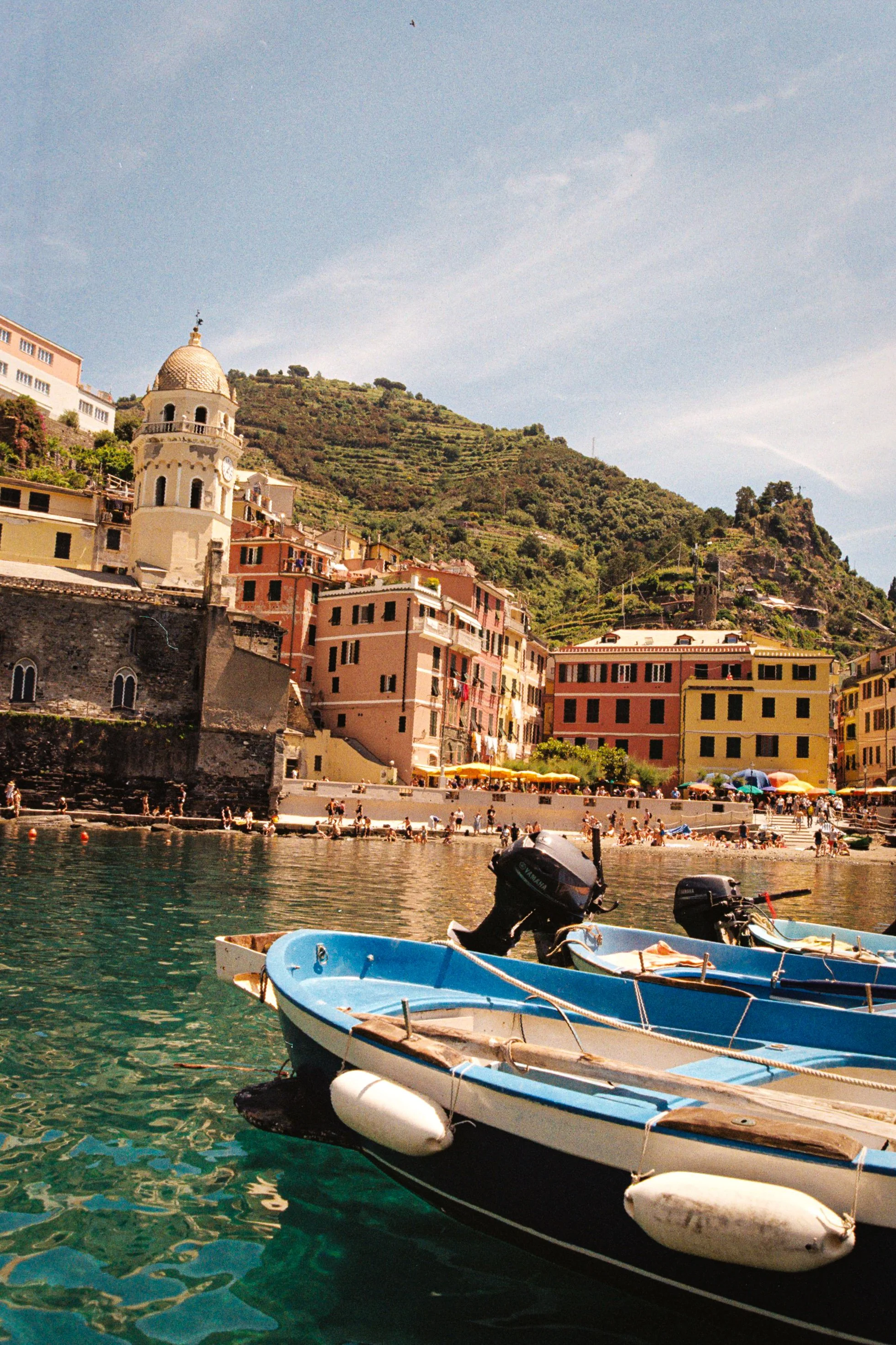 Colorful buildings on a hillside by a waterfront with boats in the water, tourists on the shore, and a partly cloudy sky.