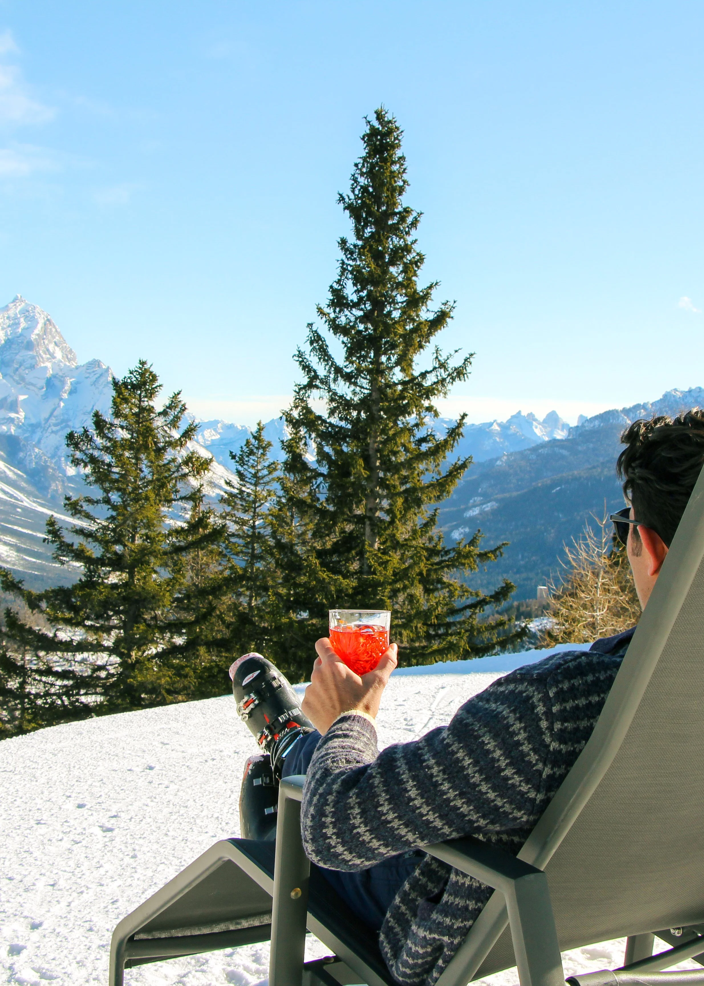 A man relaxing in a lounge chair outdoors in the snow, holding a glass of red beverage with ice, facing a scenic mountain view with snow-capped peaks and evergreen trees.