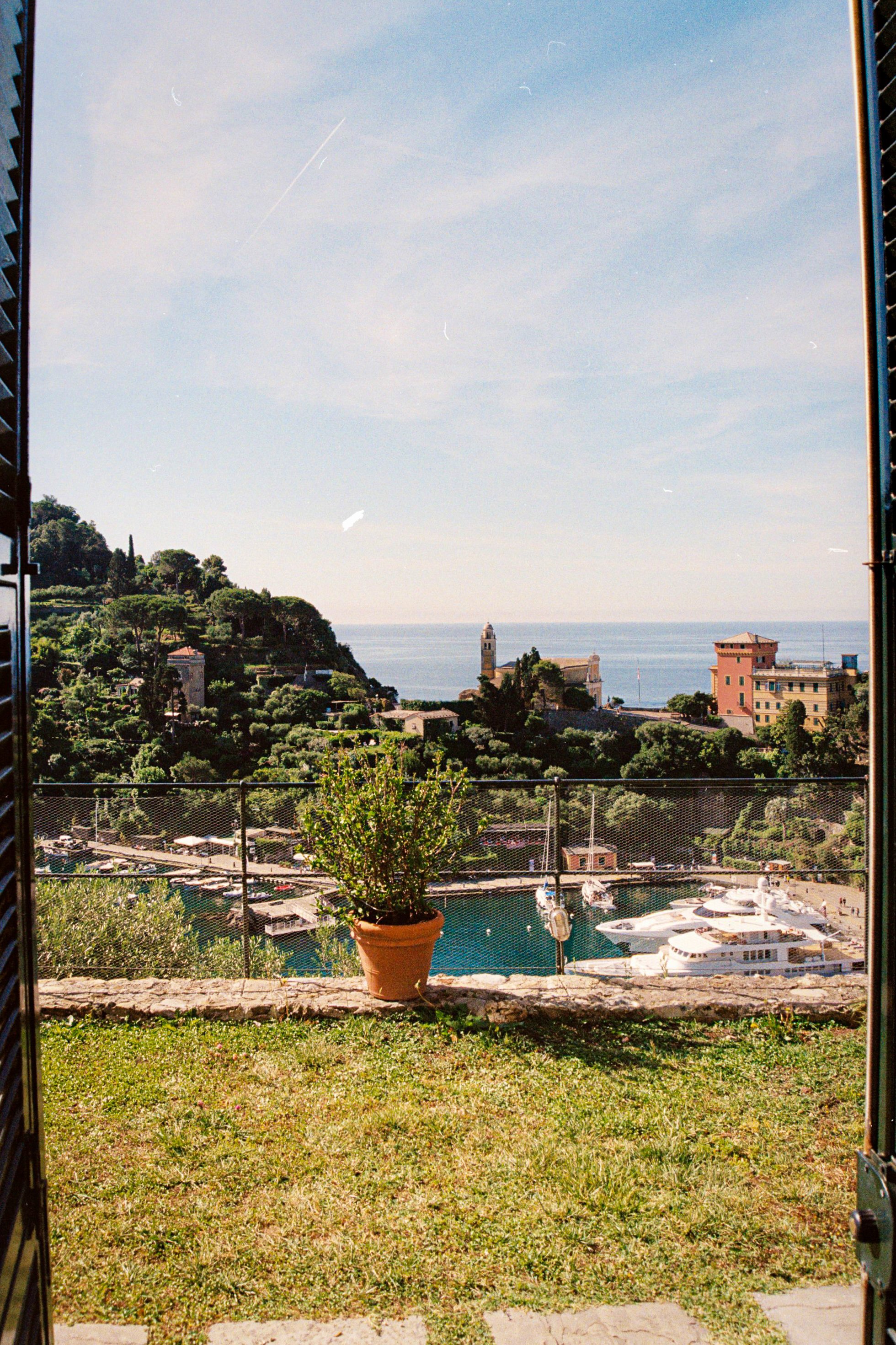 View through an open gate showing a terracotta pot with green shrubbery in a garden, overlooking a coastal landscape with hillside houses, a waterway with boats, and the sea in the background.