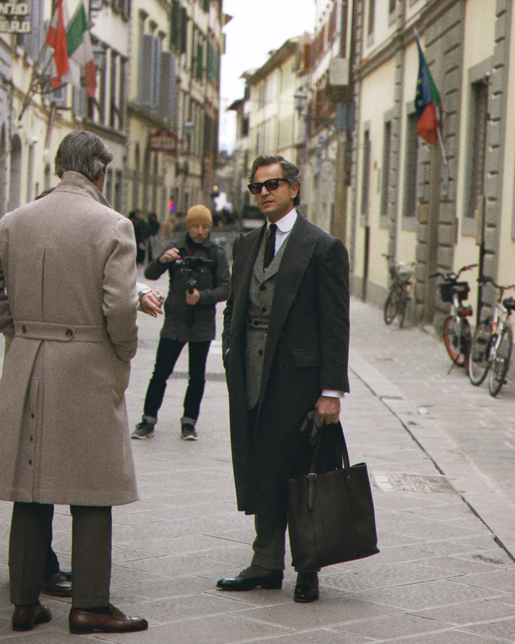 Two men are engaged in a conversation on a street with historic buildings. One is in a beige coat, and the other is in a dark coat with glasses, holding a black bag and pen. There are bicycles and a person with a camera in the background.