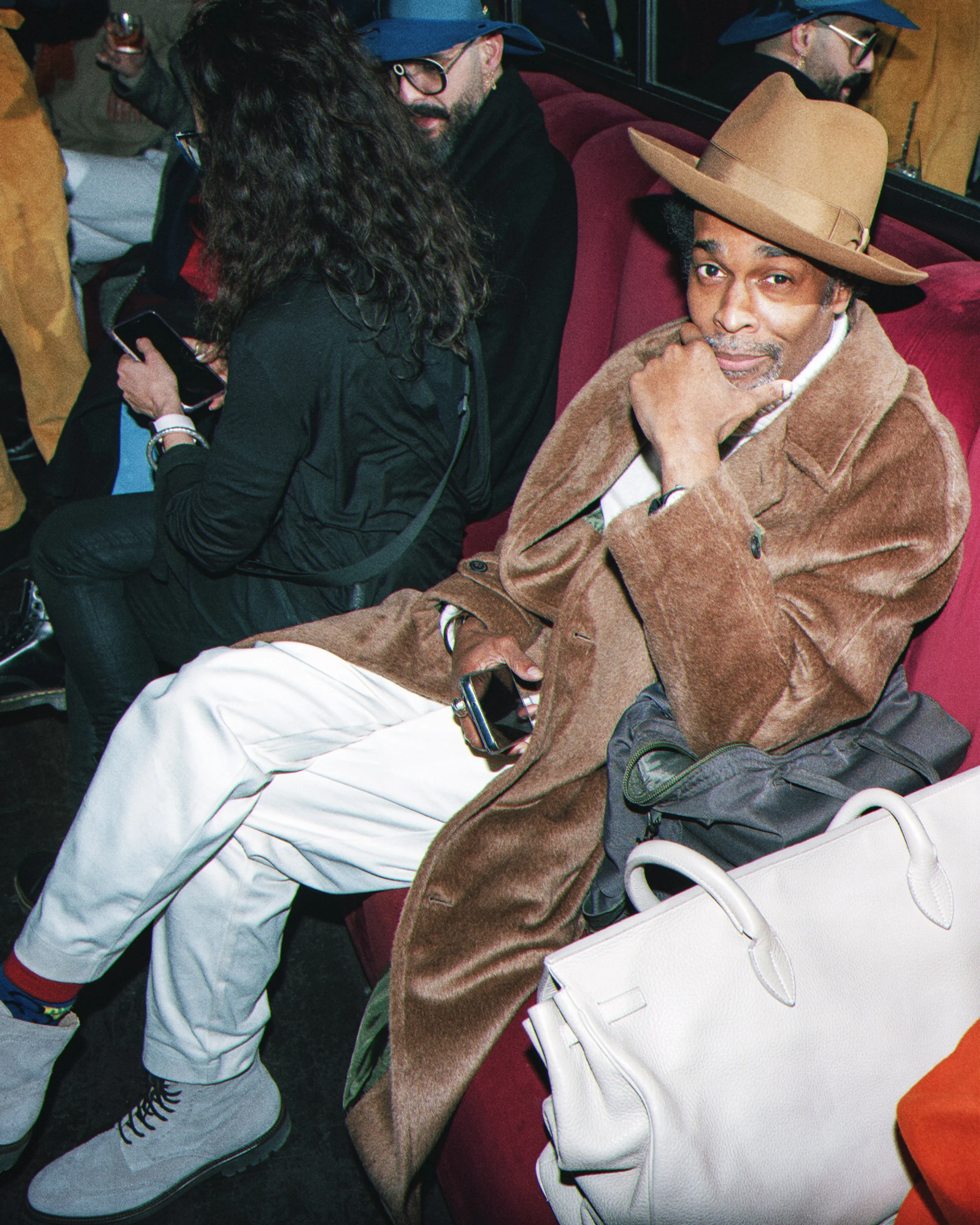 Two men and a woman sitting on a red velvet bench at a social gathering or event. One man is older, wearing a wide-brimmed tan hat, a brown coat, and has a white handbag near him. The other man is wearing a blue hat, glasses, and a dark hoodie. The w