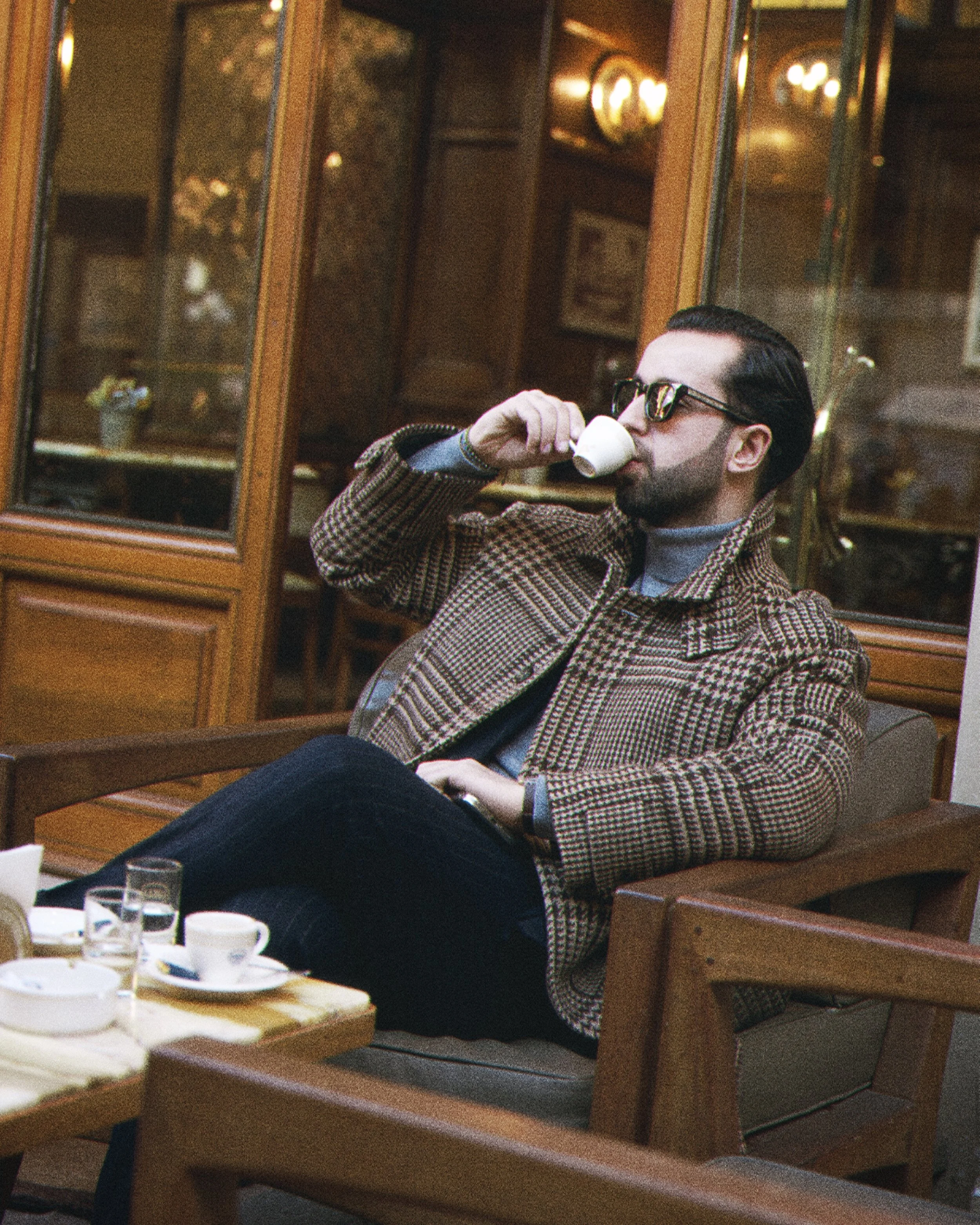 A man with dark hair, wearing glasses and a checkered brown blazer, is sitting at a table in a fancy restaurant, drinking from a small tea cup. The background shows wooden shelves and warm lighting.