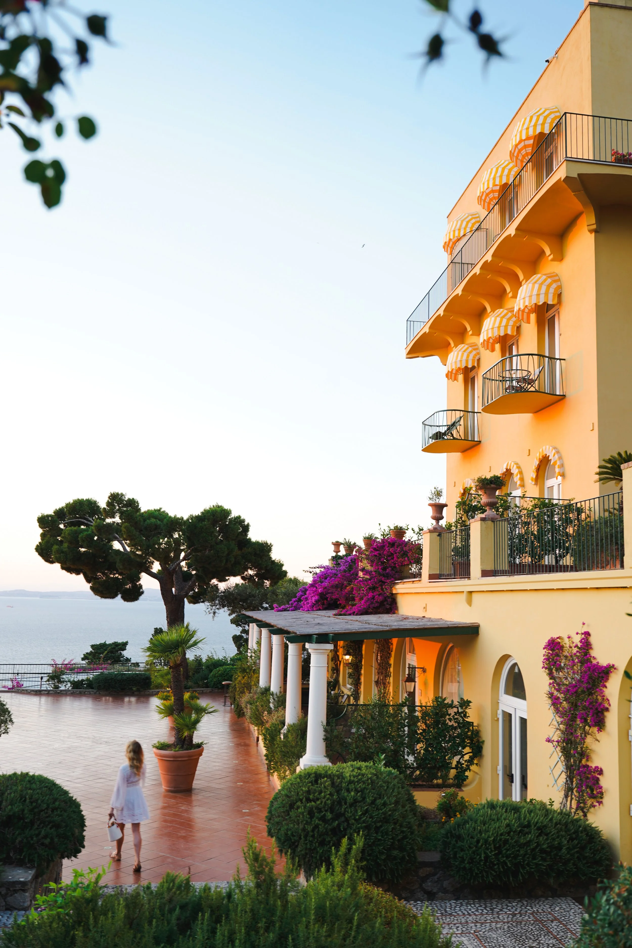 A woman walks in front of a yellow Mediterranean-style villa with balconies and striped awnings, overlooking the ocean at sunset, with lush greenery and flowering plants surrounding the area.