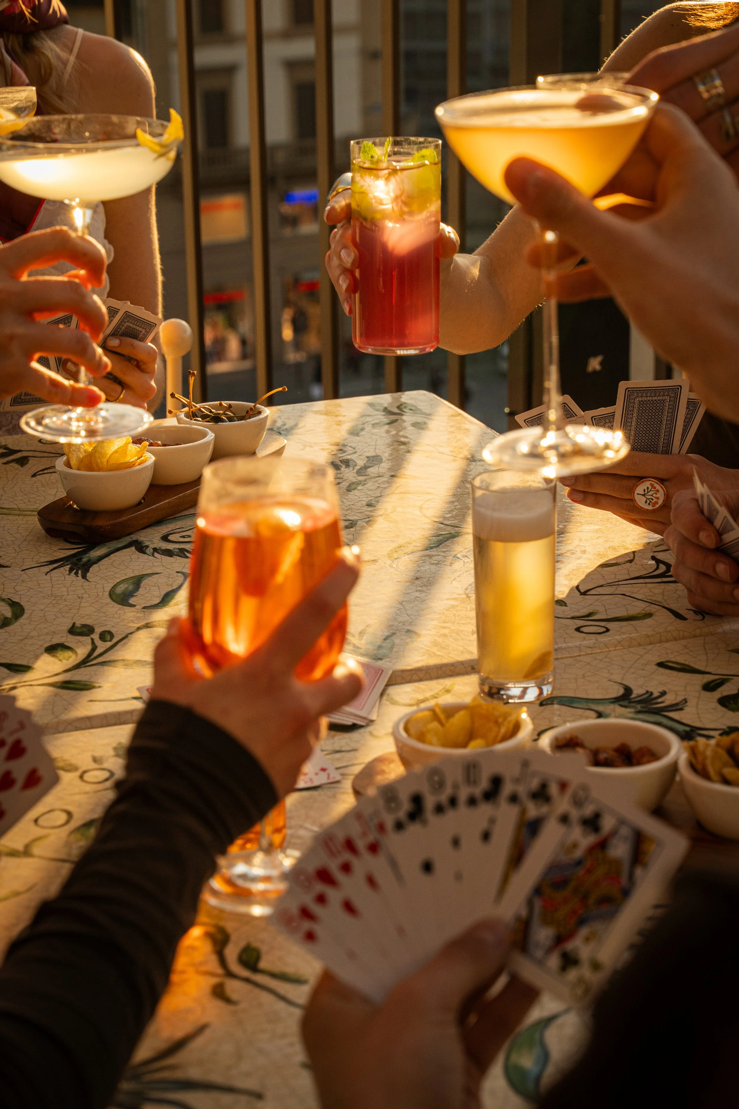 People holding cocktails and playing cards around a table on a balcony during sunset.