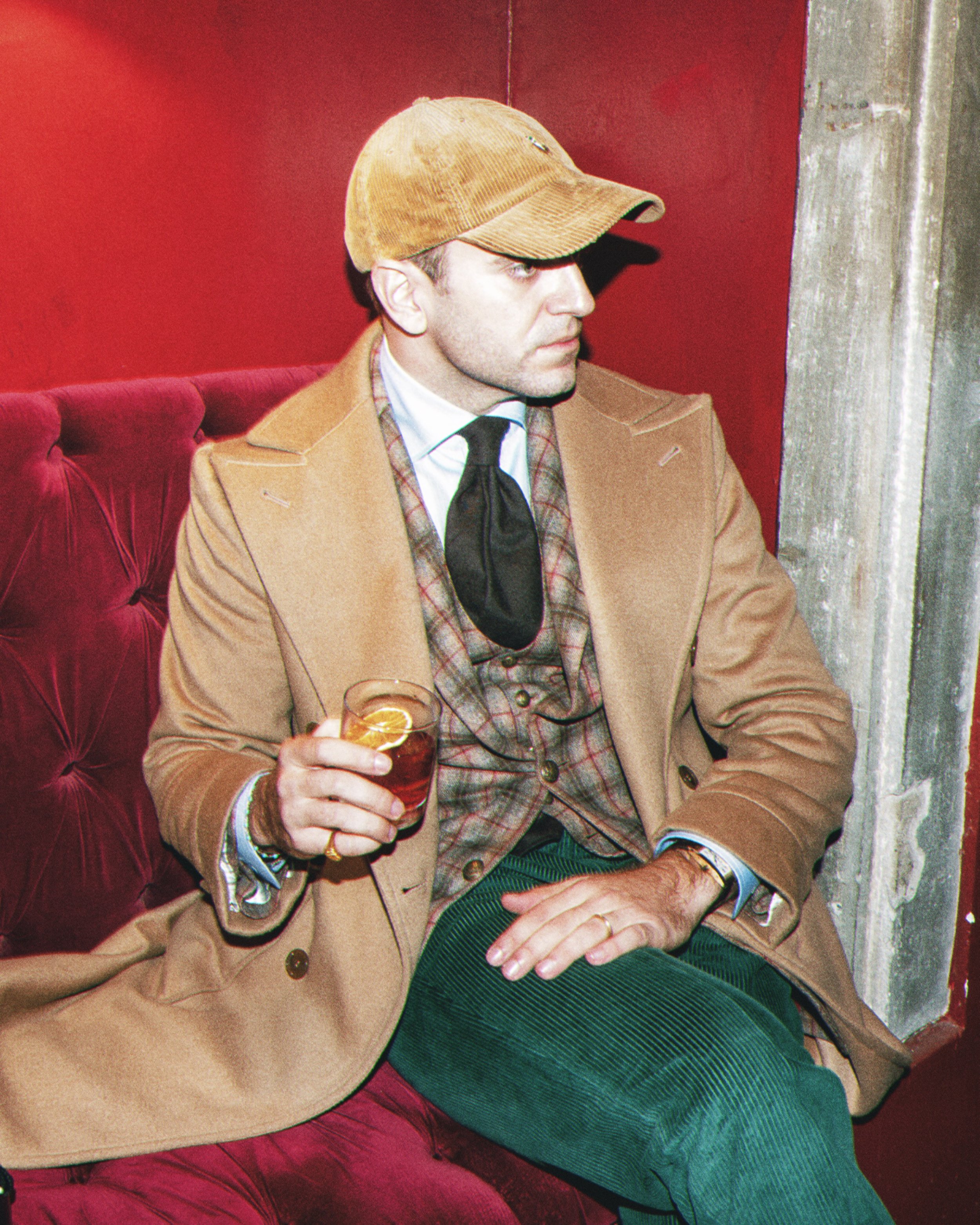 A man sitting on a red velvet bench, wearing a tan coat, a plaid vest, a white shirt, a black tie, and a tan corduroy cap. He is holding a drink with a slice of orange and red fruit in his right hand, resting his left hand on his lap. There is a gray