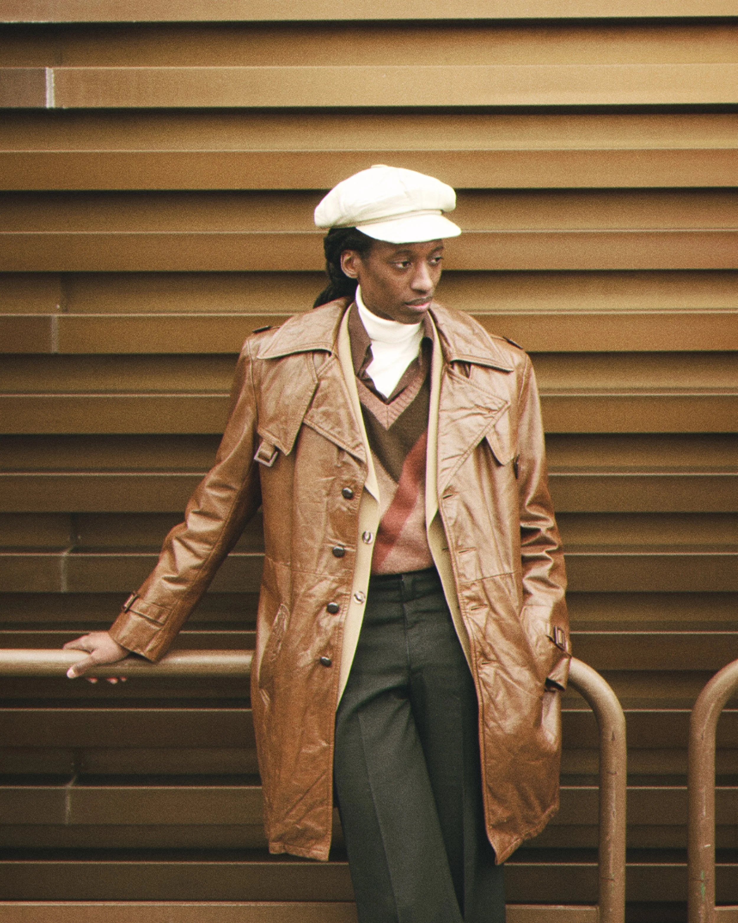 Person wearing a beige cap and brown trench coat standing against a brown metal fence.