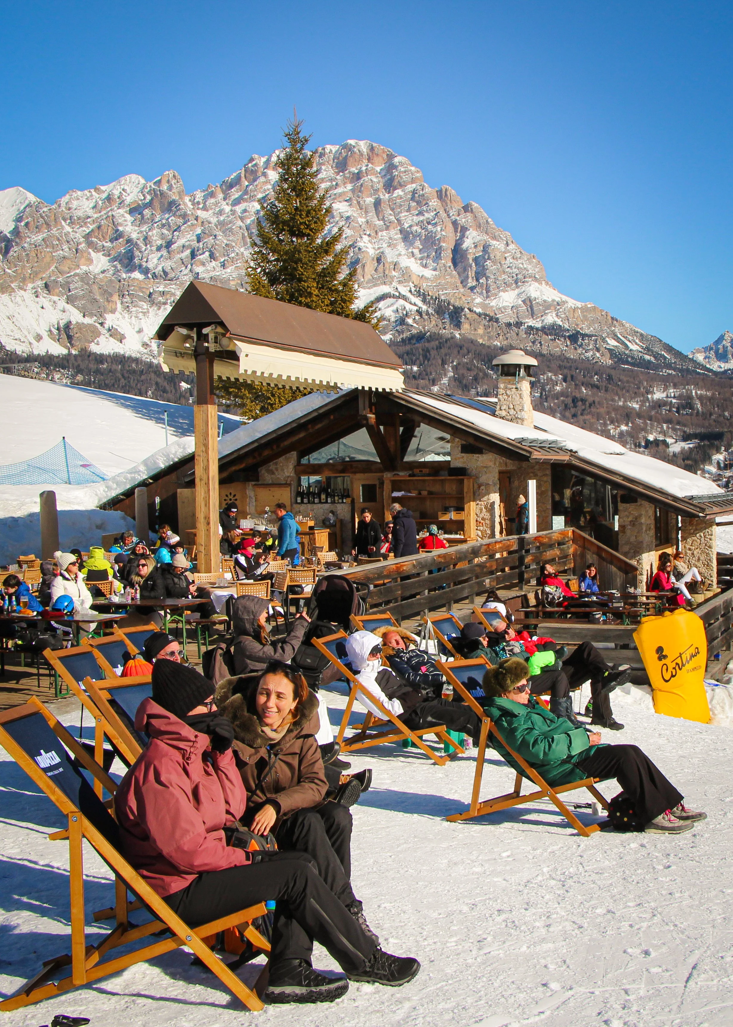 People sitting on lounge chairs outside at a ski resort, with snow-covered ground, a chalet building, a mountain with snow, and a clear blue sky in the background.