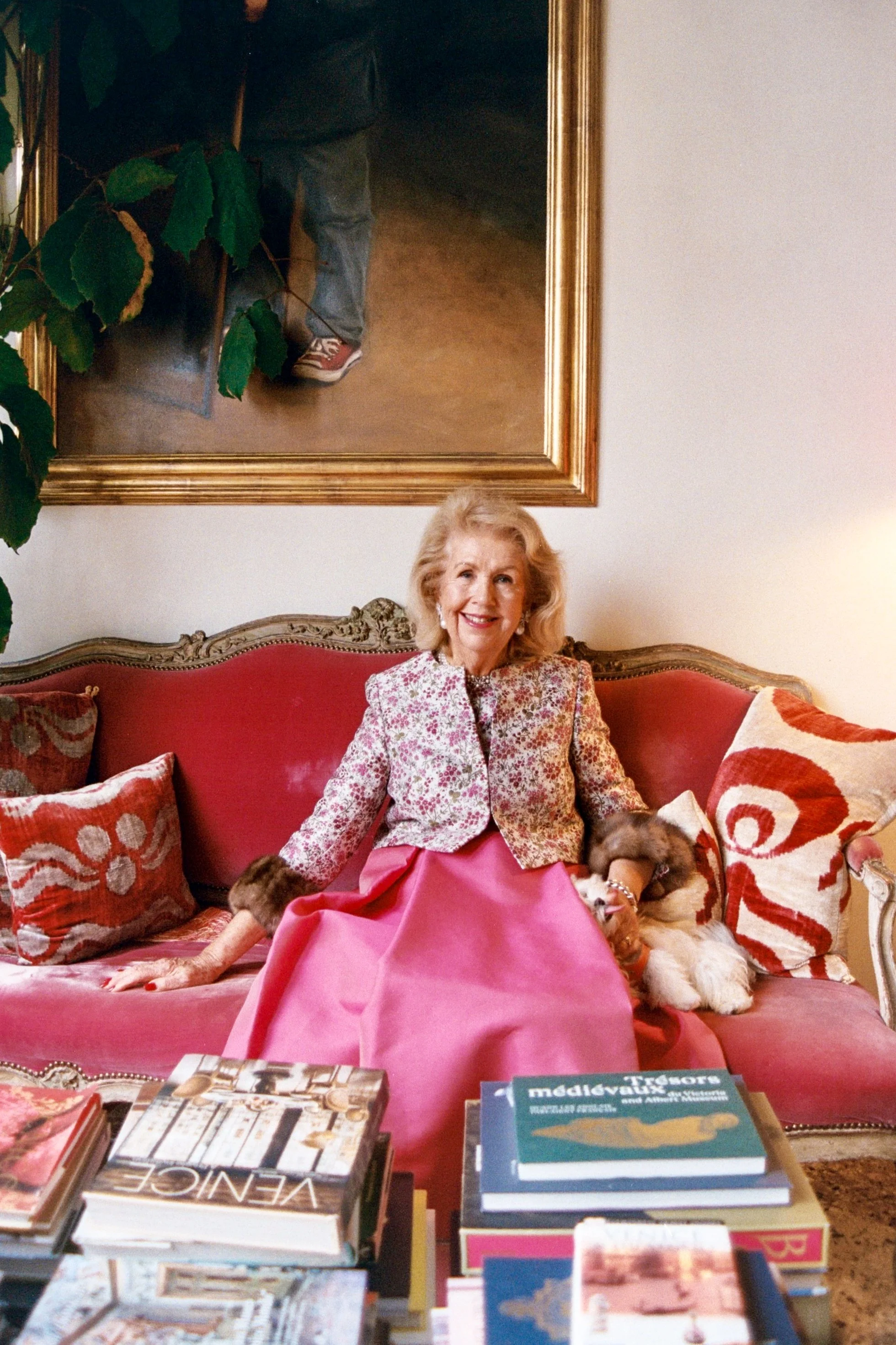 An elderly woman with blonde hair, wearing a floral jacket and pink skirt, sitting on a red velvet sofa with ornate wooden trim, smiling, with a small dog beside her. There are books on a table in front of her and abstract patterned pillows behind he
