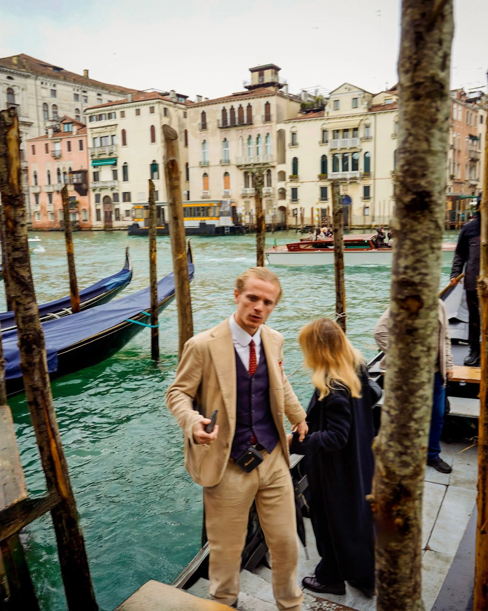 A man in beige suit and a woman with blonde hair talking near a canal with gondolas in Venice, Italy. Multiple boats and colorful buildings are visible in the background.