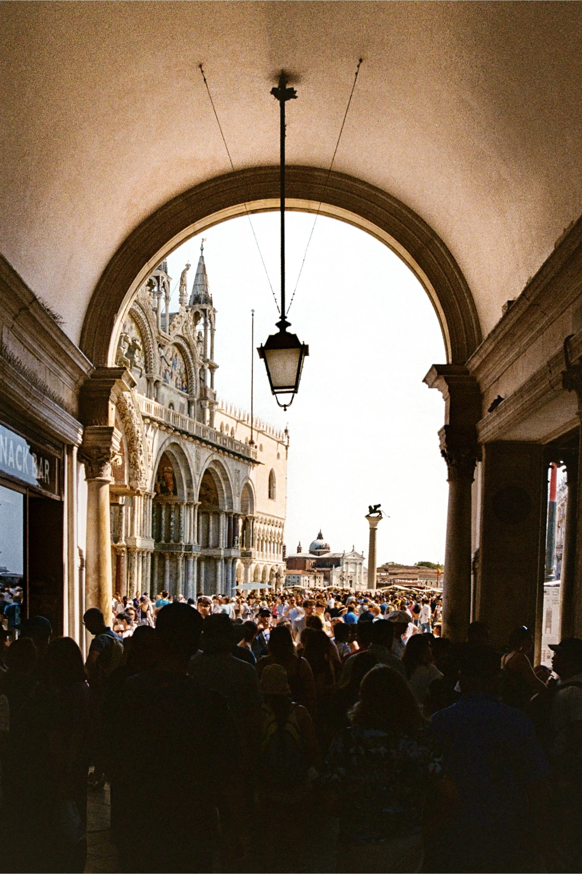 View of St. Mark's Square in Venice, Italy, seen from under an archway with a hanging lantern. The square is crowded with people, and the Doge's Palace's ornate façade is visible on the left. In the background, there are historic buildings and a colu