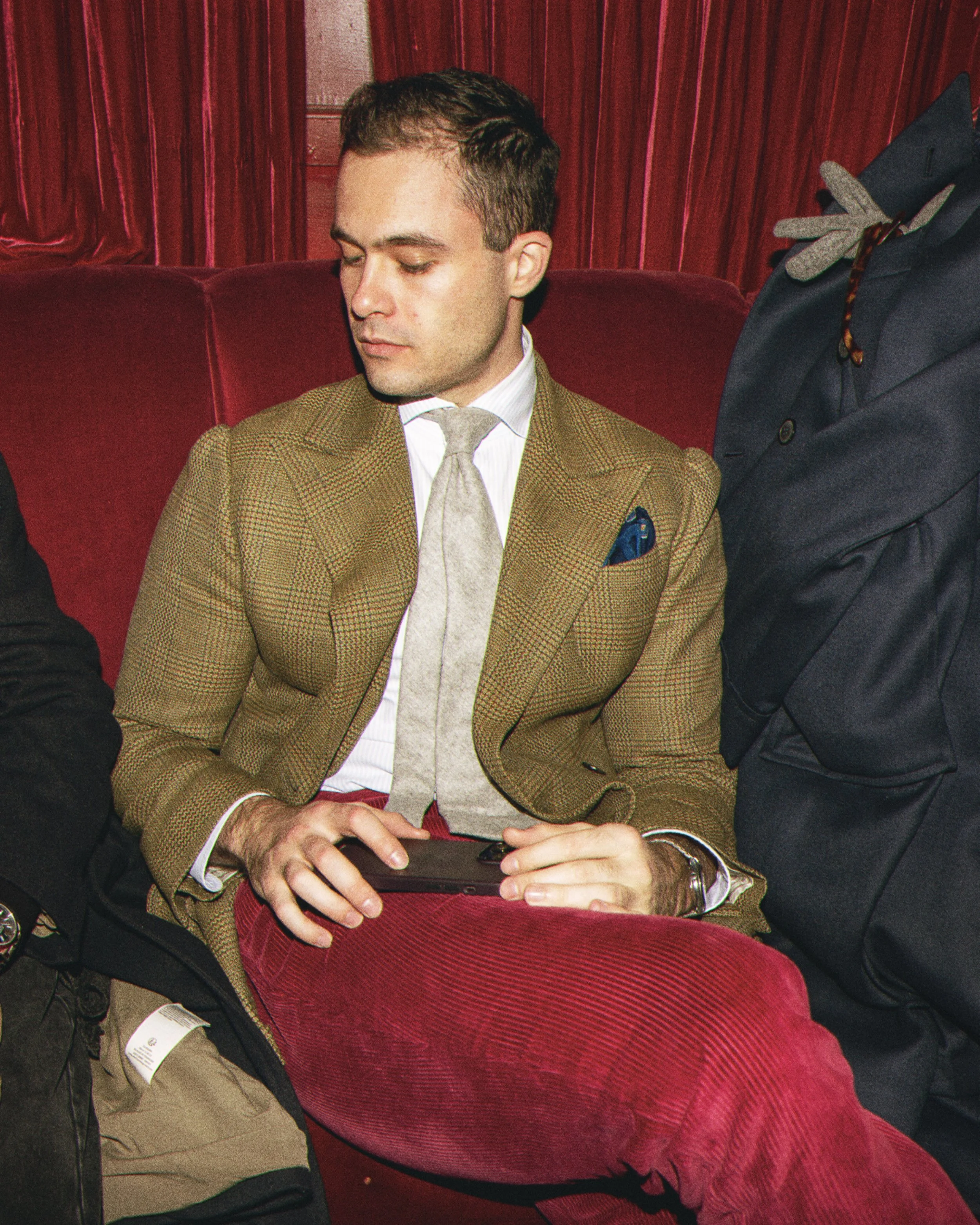 A man in a brown blazer and gray tie sitting with his eyes closed, holding a smartphone, with a person in a black suit and red sweater sitting beside him on a red cushioned seat, in front of red curtains.