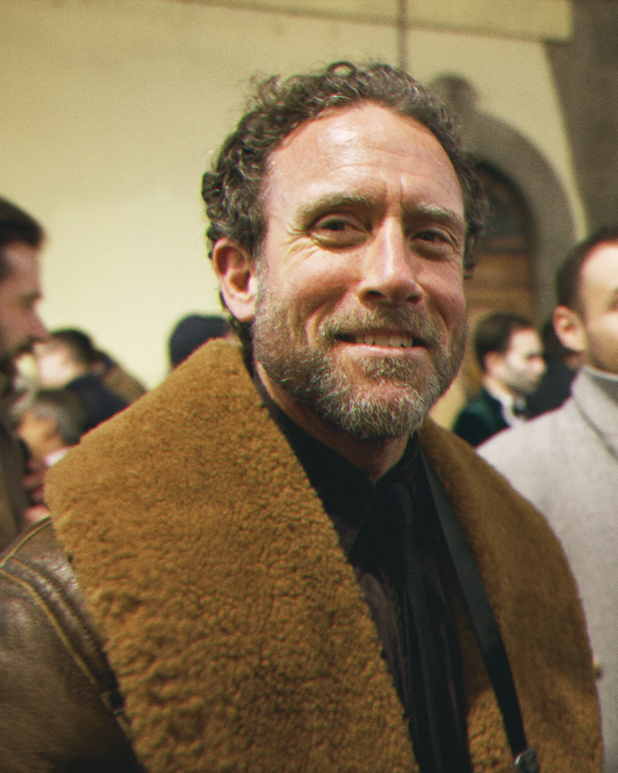Close-up of a smiling man with curly hair and a beard, wearing a brown coat, in a room with other people in the background.