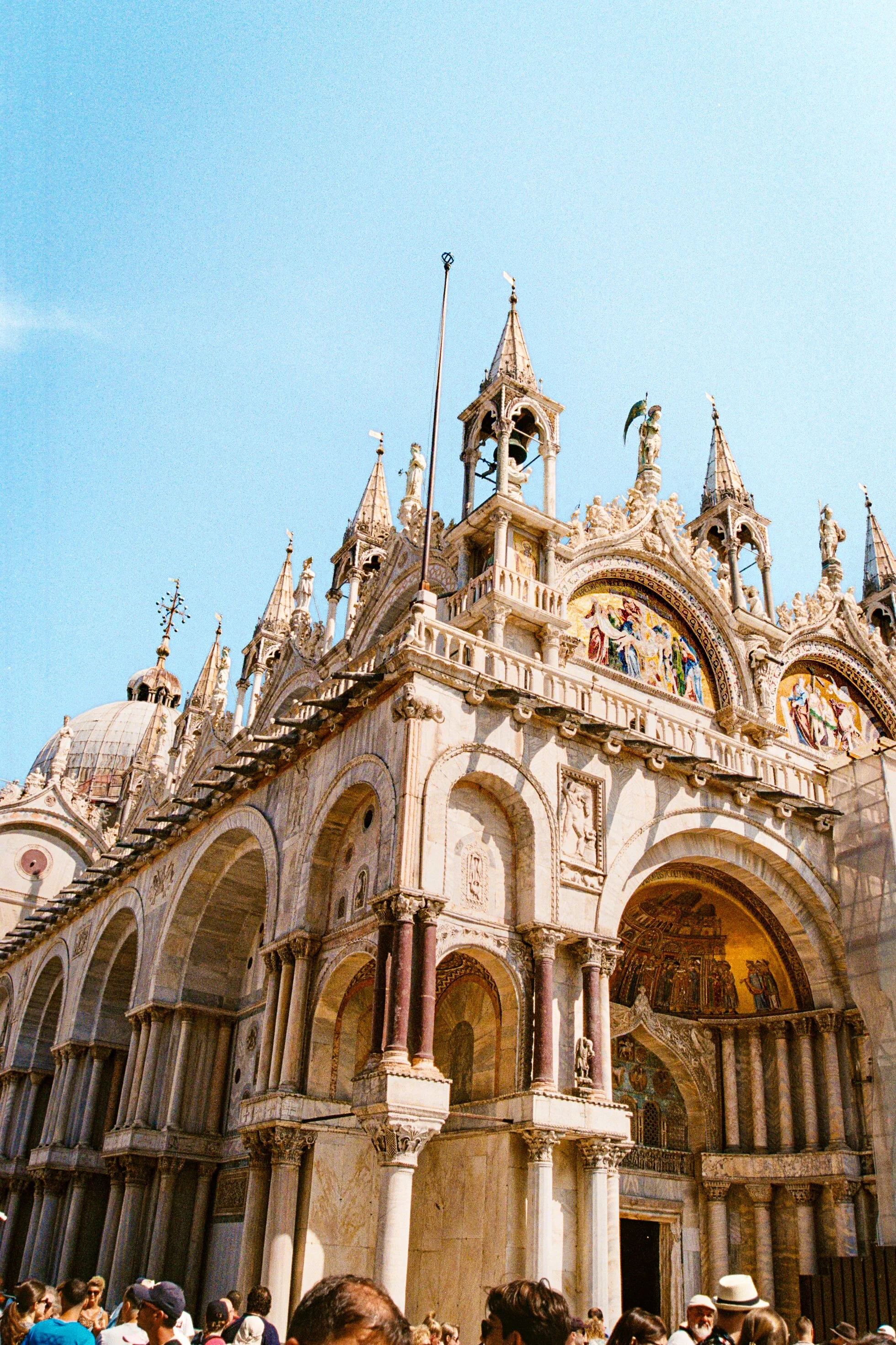 Detailed view of a historic European cathedral with ornate architecture, domes, spires, and statues, likely Saint Mark's Basilica in Venice, Italy, with tourists at the base.