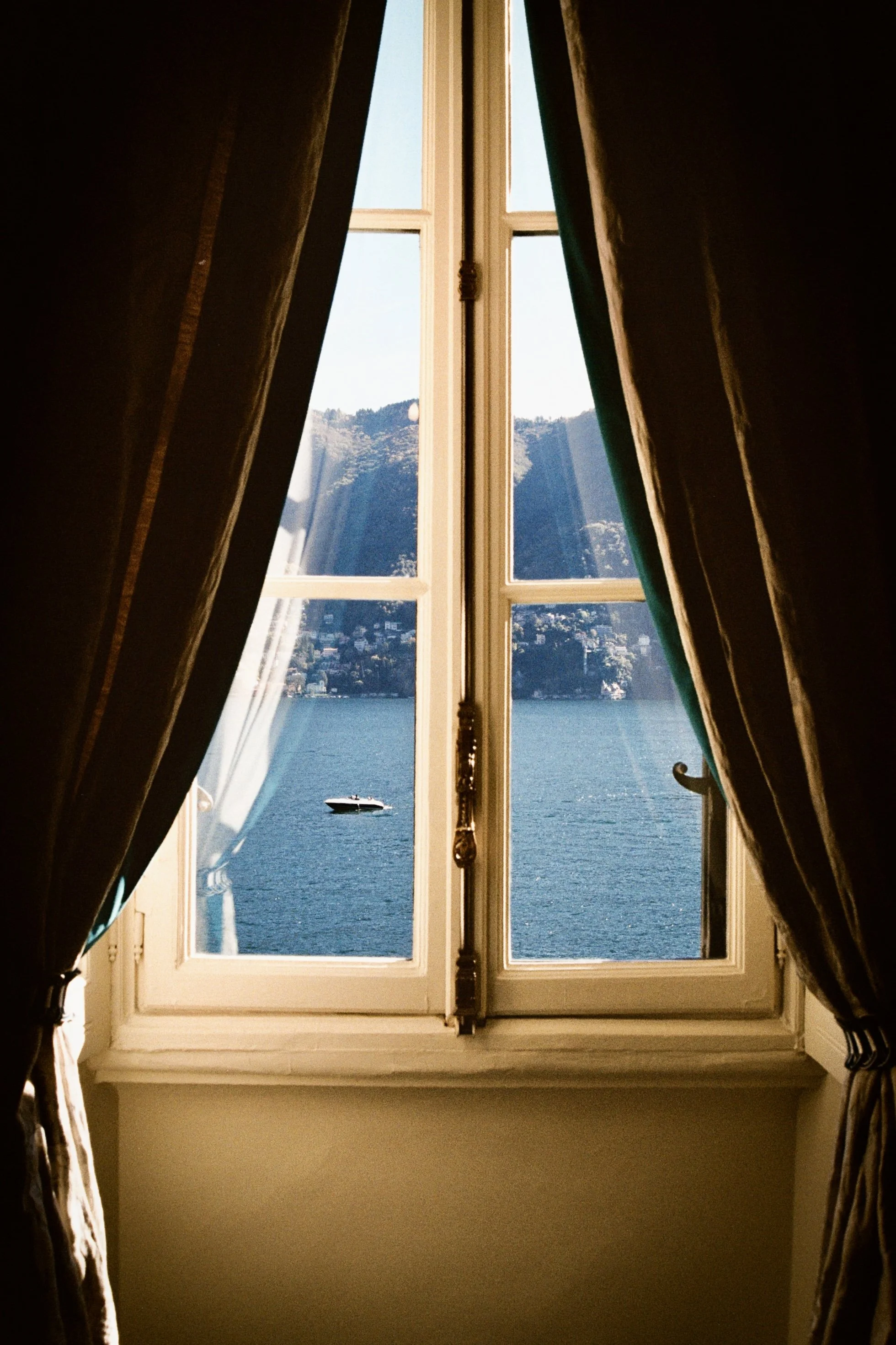 A view through an open window with curtains, showing a body of water, a boat, and mountains in the background.