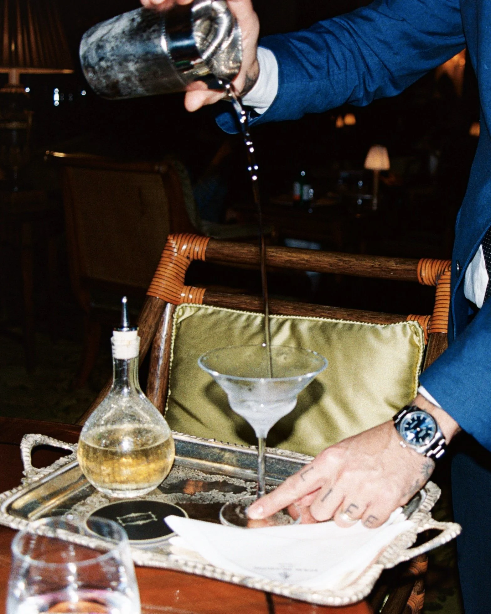A man in a blue jacket is pouring a drink into a martini glass on a tray, with a bottle and a black jar on the tray, and a person's hand with rings pointing to a dish on the tray.
