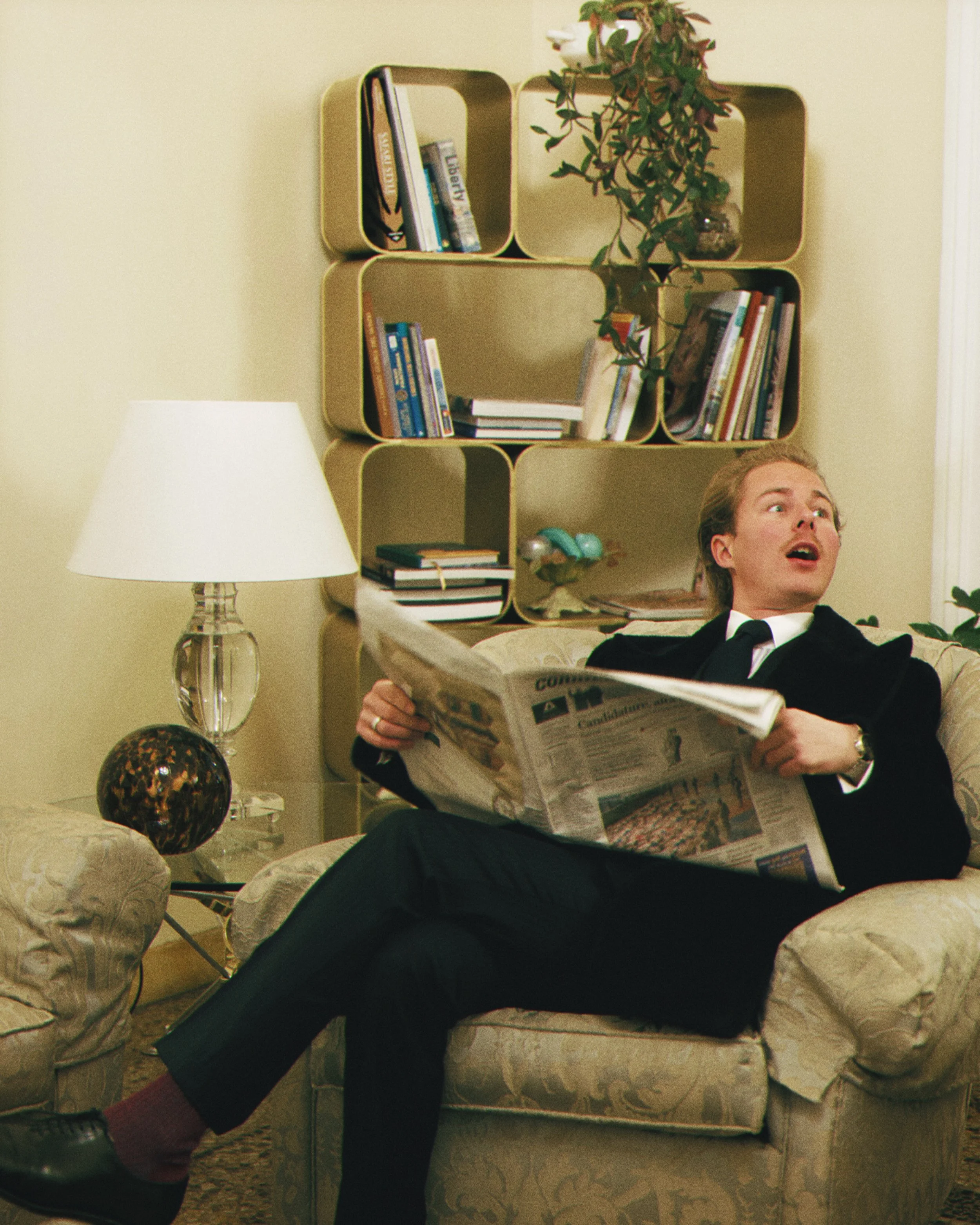 A man sitting on a floral-patterned armchair in a living room, reading a newspaper. The room has a beige wall, a white table lamp with a glass base, a decorative glass sphere, a wall-mounted shelf with books and a plant, and various decorative items 