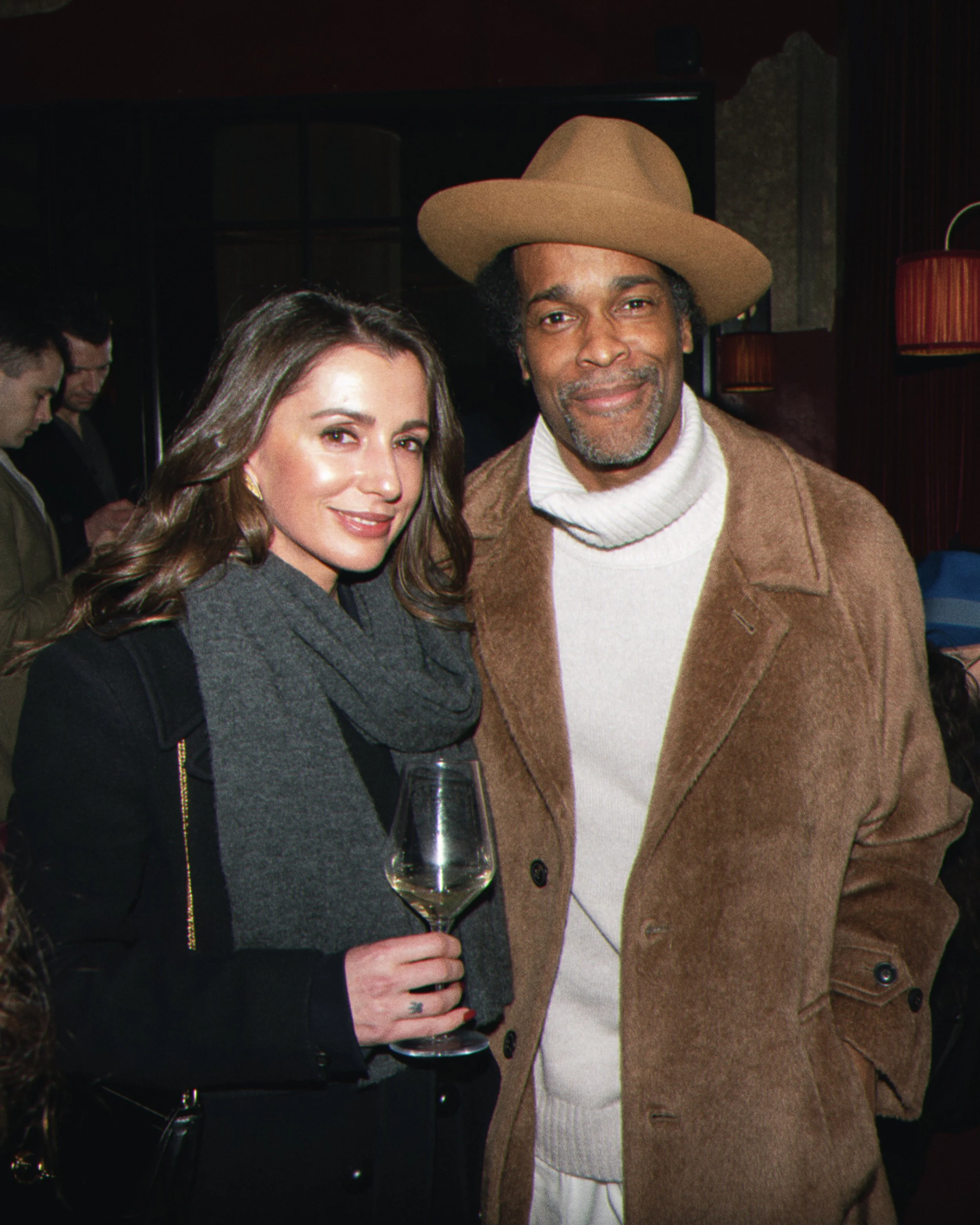A woman with long wavy hair, wearing a dark gray coat and gold earrings, and a man with a beard, wearing a tan hat, a brown coat, and a white turtleneck sweater, smiling at the camera and holding a glass of white wine in a dimly lit setting.