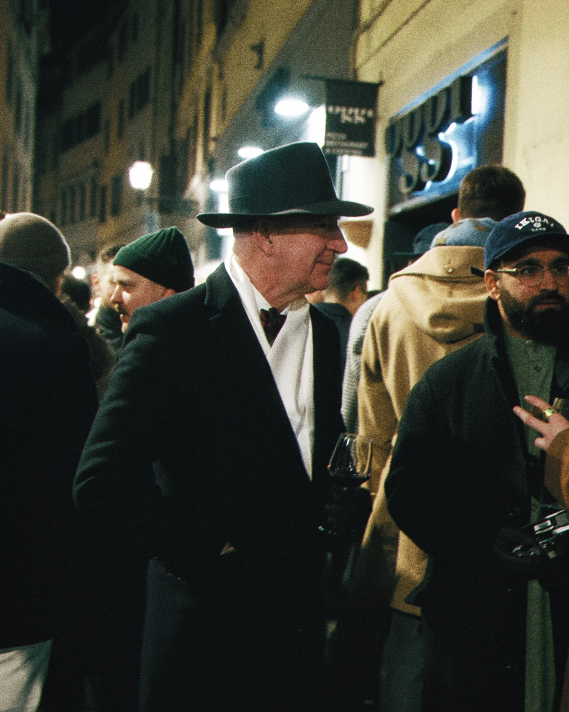 People standing outdoors at night, including a man in a black hat and suit, a man in a green beanie, and a person in a tan coat with a hoodie, in front of a building with illuminated signs.