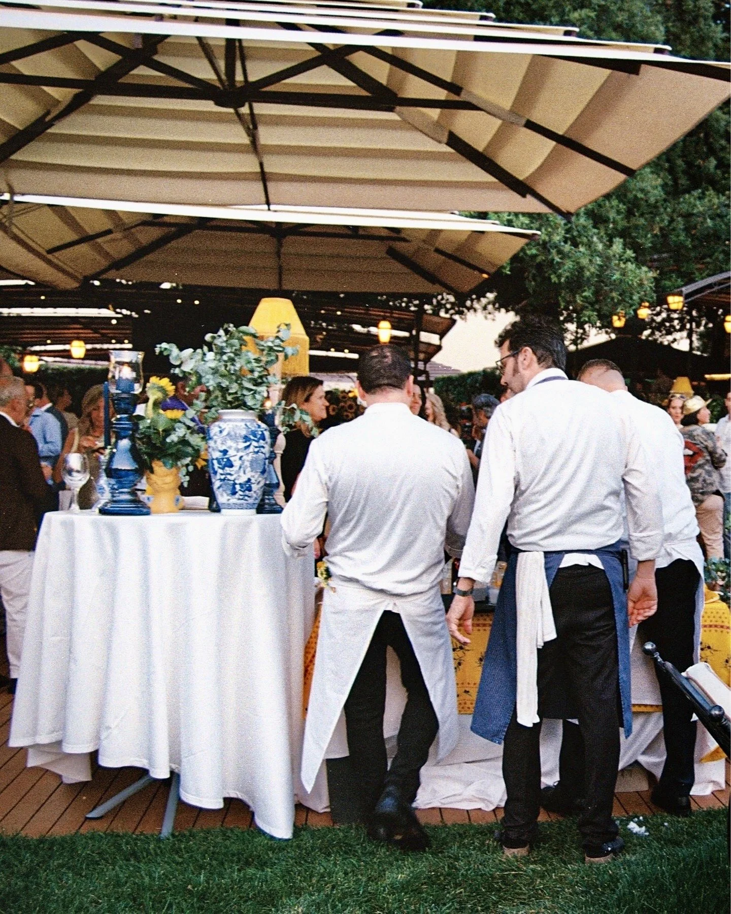 People gathered at an outdoor event under large beige umbrellas, with a table decorated with blue and white vases and flowers, and two men in white shirts with aprons facing away from the camera.