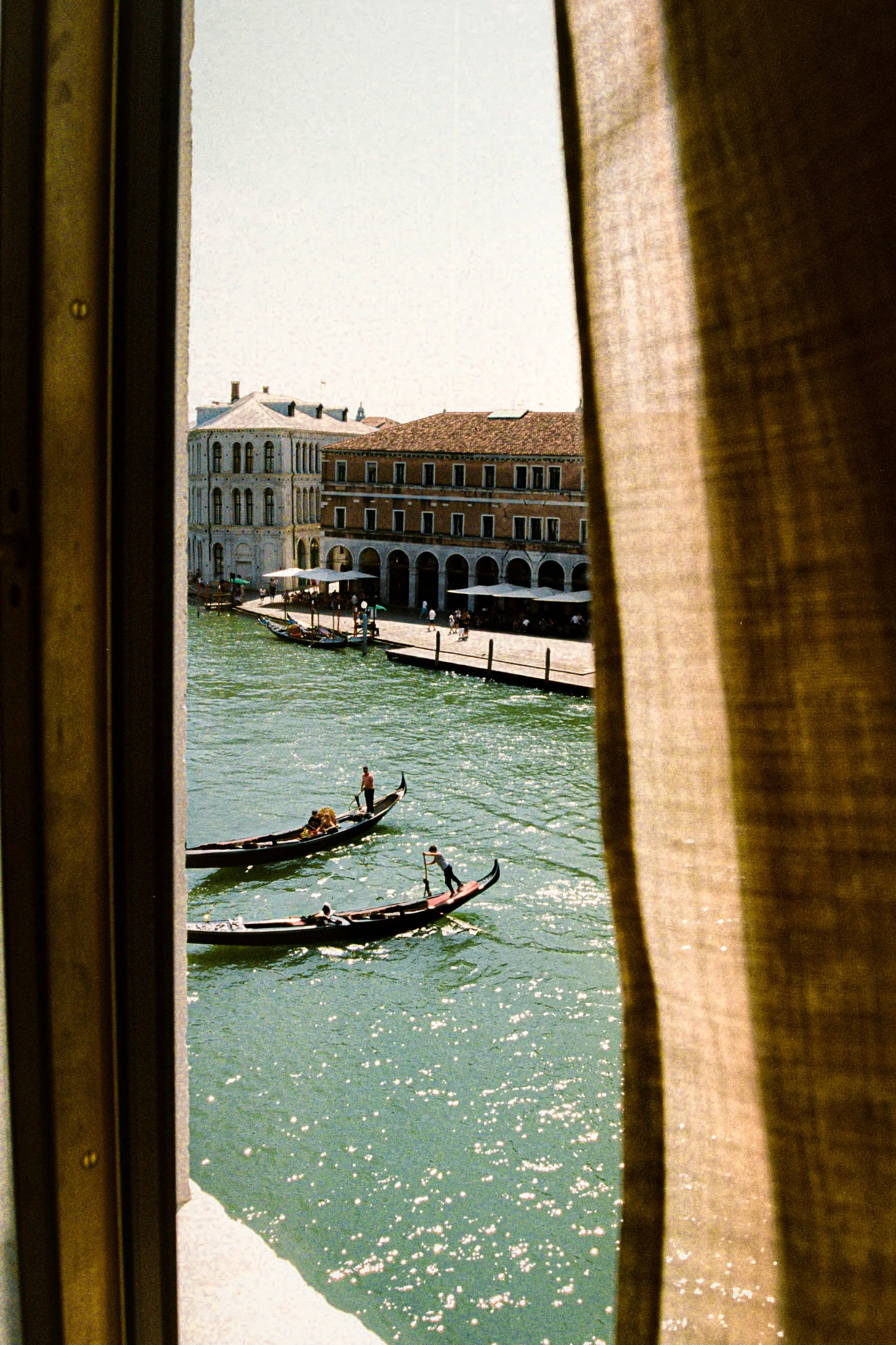 View of the Grand Canal in Venice through a window with brown curtains, showing gondolas and historic buildings along the water.