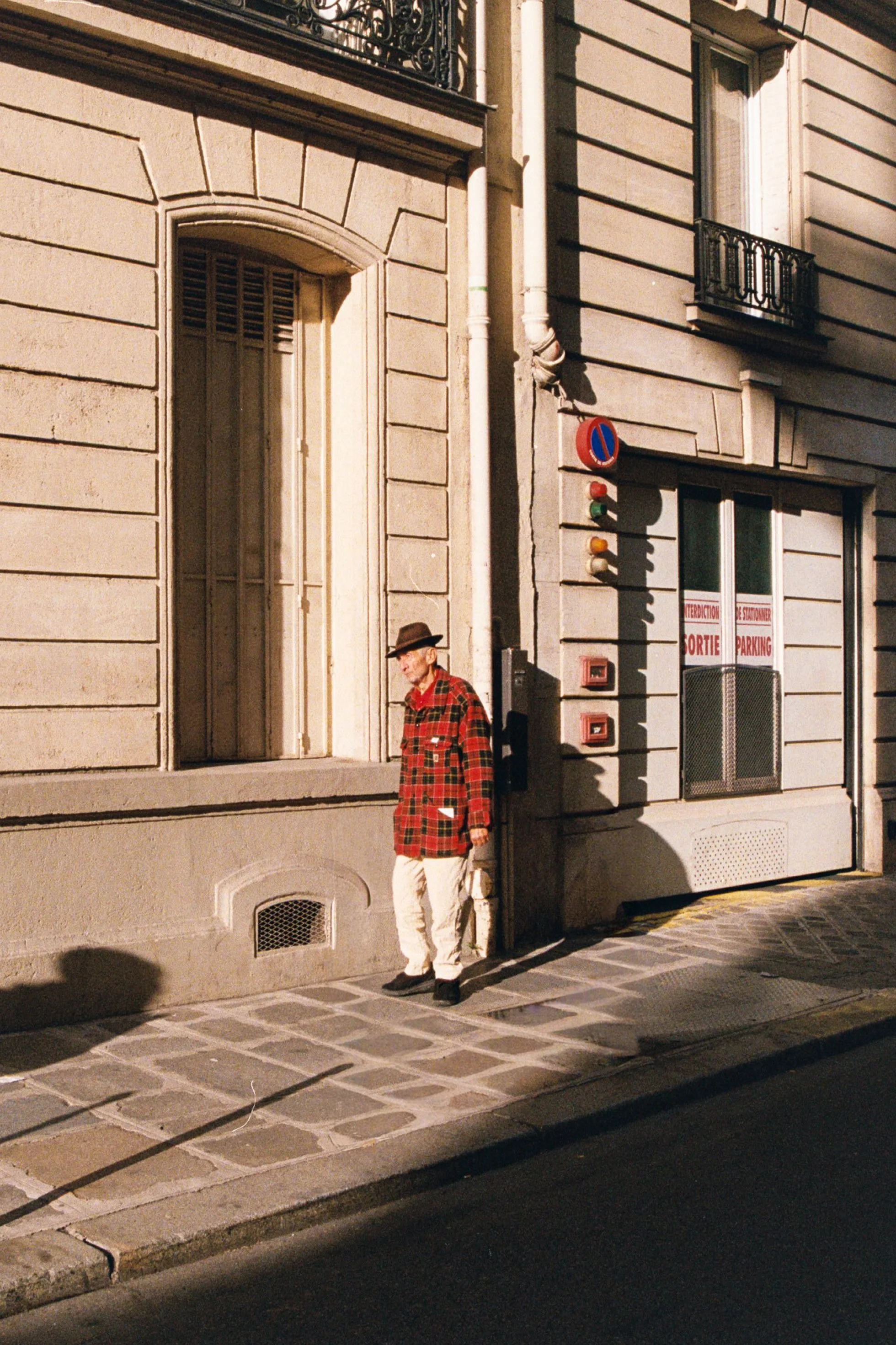 An elderly man wearing a red plaid jacket, white pants, brown hat, and glasses standing on a city sidewalk next to a beige building with barred windows and a set of traffic lights.