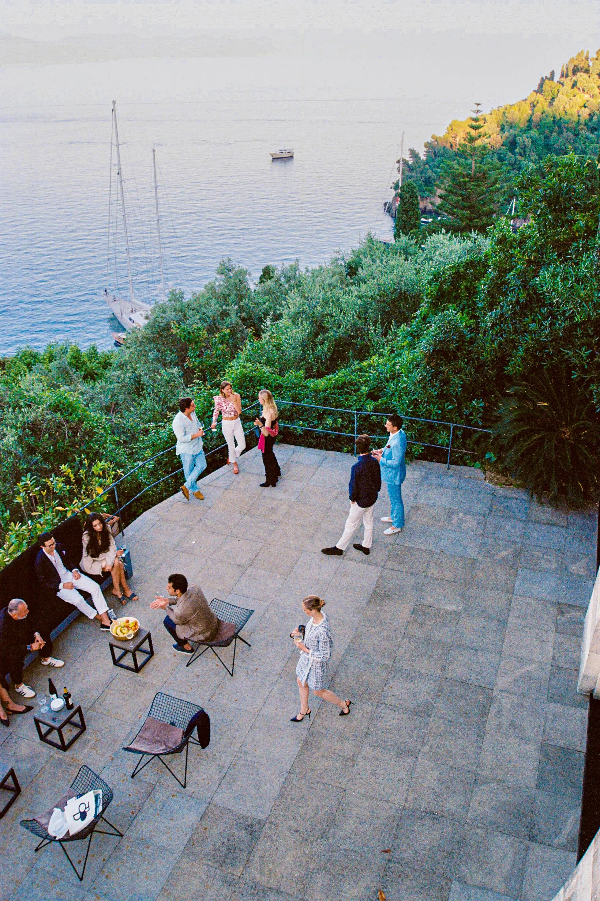 People socializing on a terrace with a scenic view of the ocean, trees, sailboats, and hills in the background.